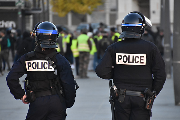 Two police officers wearing helmets and tactical gear stand with backs to the camera, observing a crowd of people wearing neon yellow vests at an outdoor event or protest.