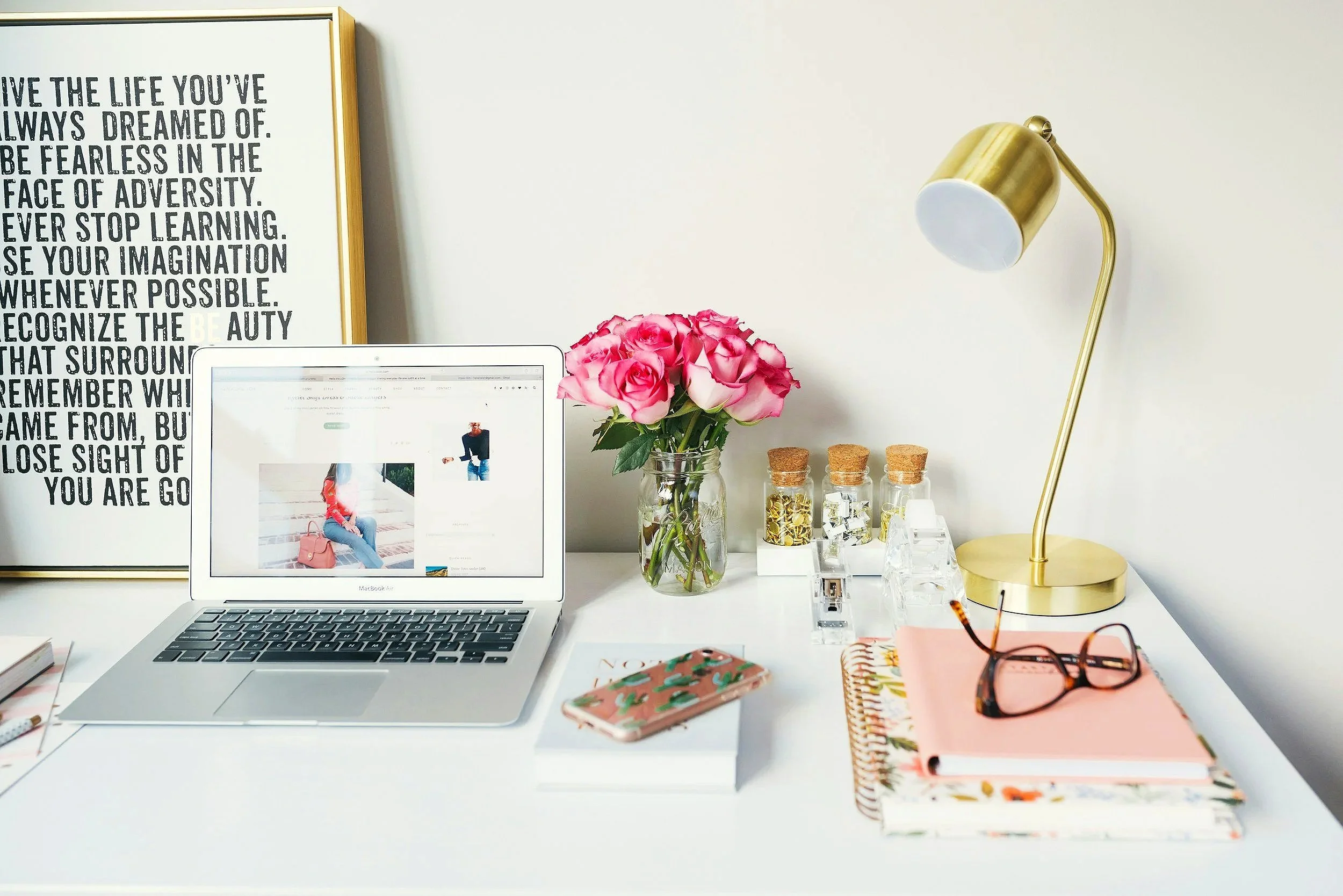 A white desk with a laptop, pink roses in a glass vase, notebooks, a pink phone case, a pair of glasses, and a gold desk lamp. There is a framed quote on the wall behind the desk.