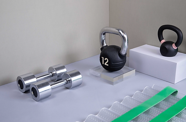 Dumbbells, a kettlebell, a kettlebell with the number 12 on it, and a kettlebell with a pink accent, all arranged on a white surface against a neutral wall.