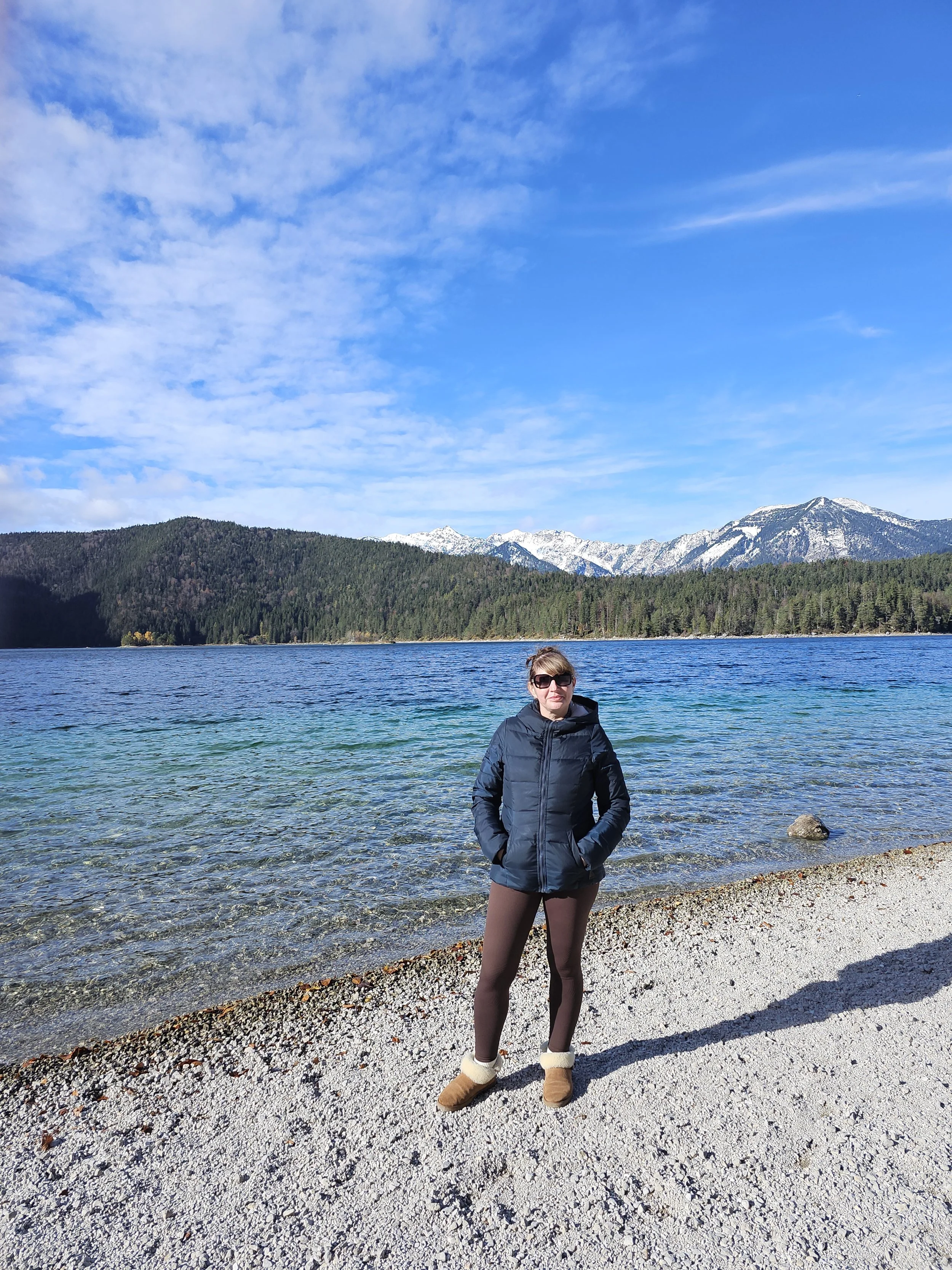 A woman in sunglasses and a black jacket stands on a rocky beach with a mountain lake and snow-capped mountains in the background.