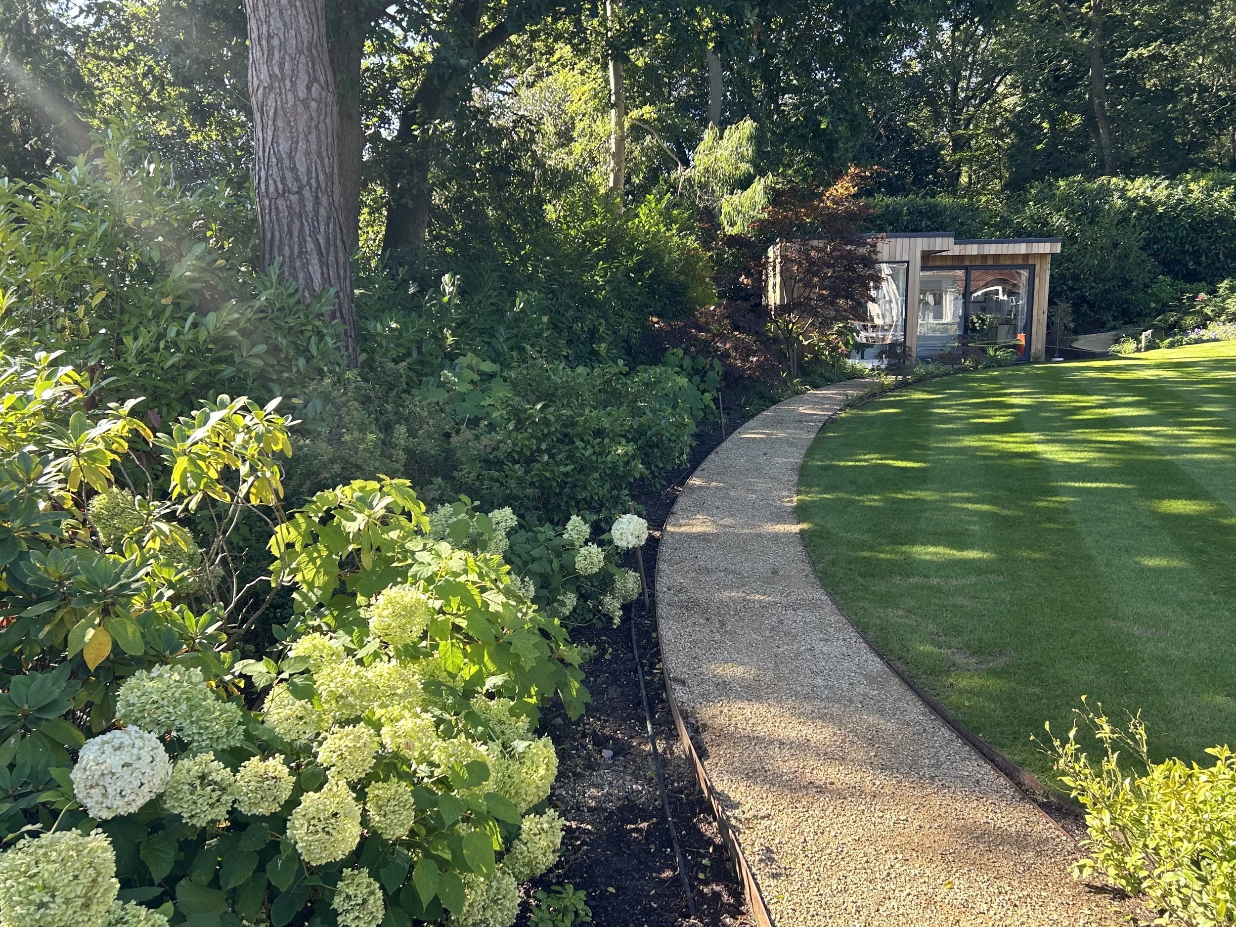 A curved concrete pathway running through a well-maintained lawn and garden with various green shrubs and trees, leading to a small modern glass building in the background.
