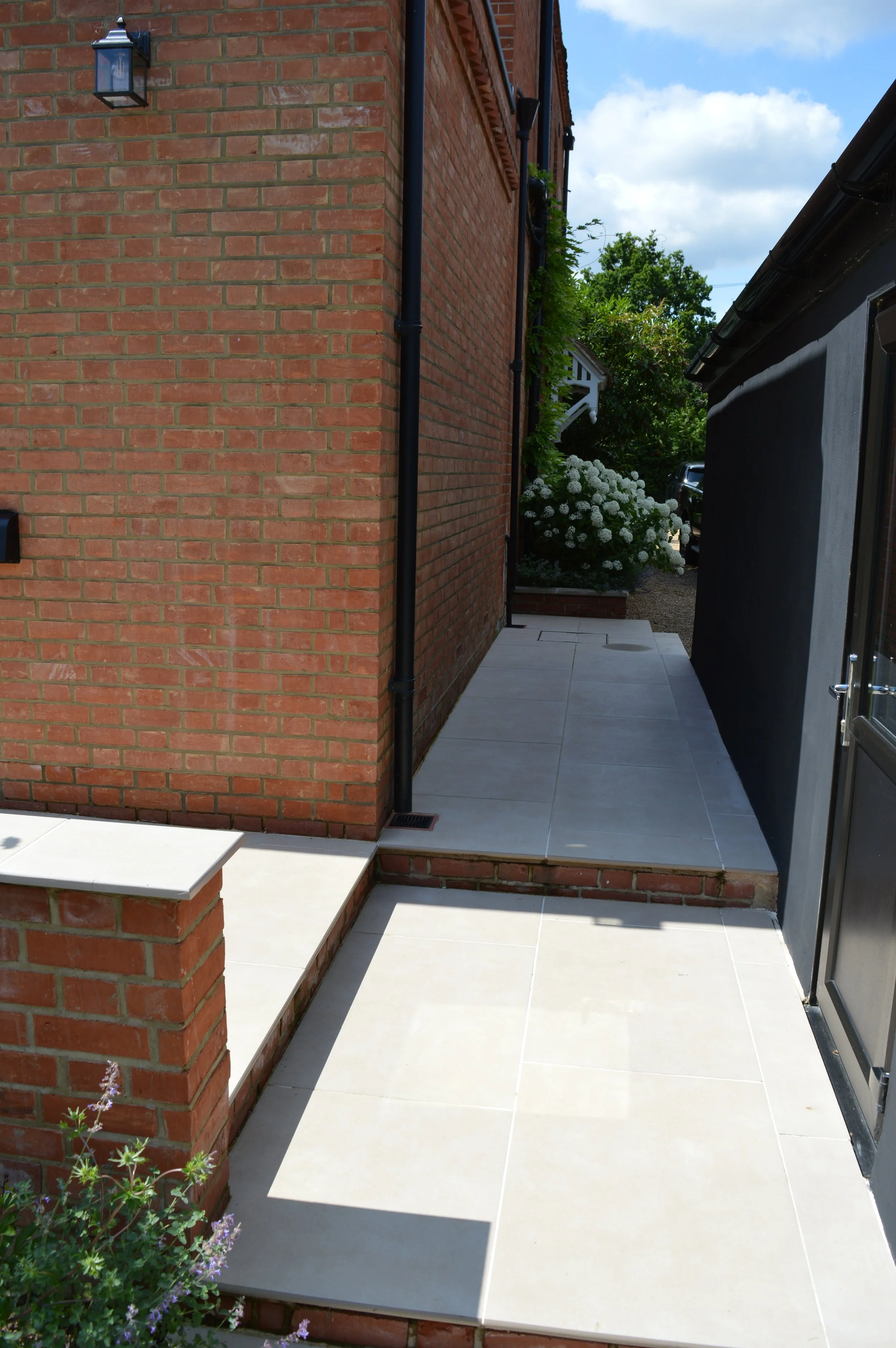 Side view of a house with a brick wall, outdoor paving, a black drainpipe, plants, and a flowering bush under a partly cloudy sky.