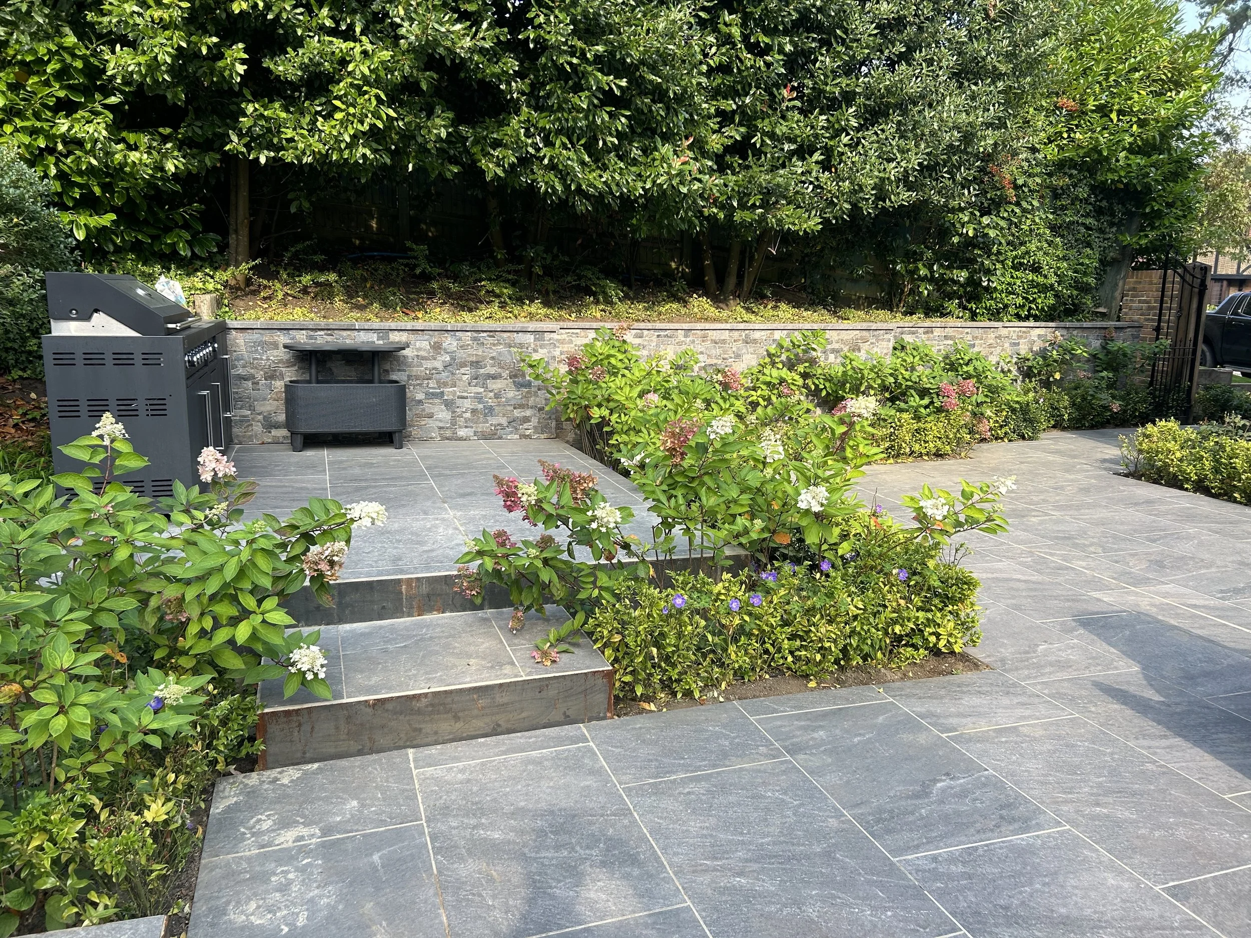 Backyard patio with gray stone tiles, a black grill, a small black table, and flowering bushes with pink, white, and purple flowers, bordered by a stone wall and green trees.