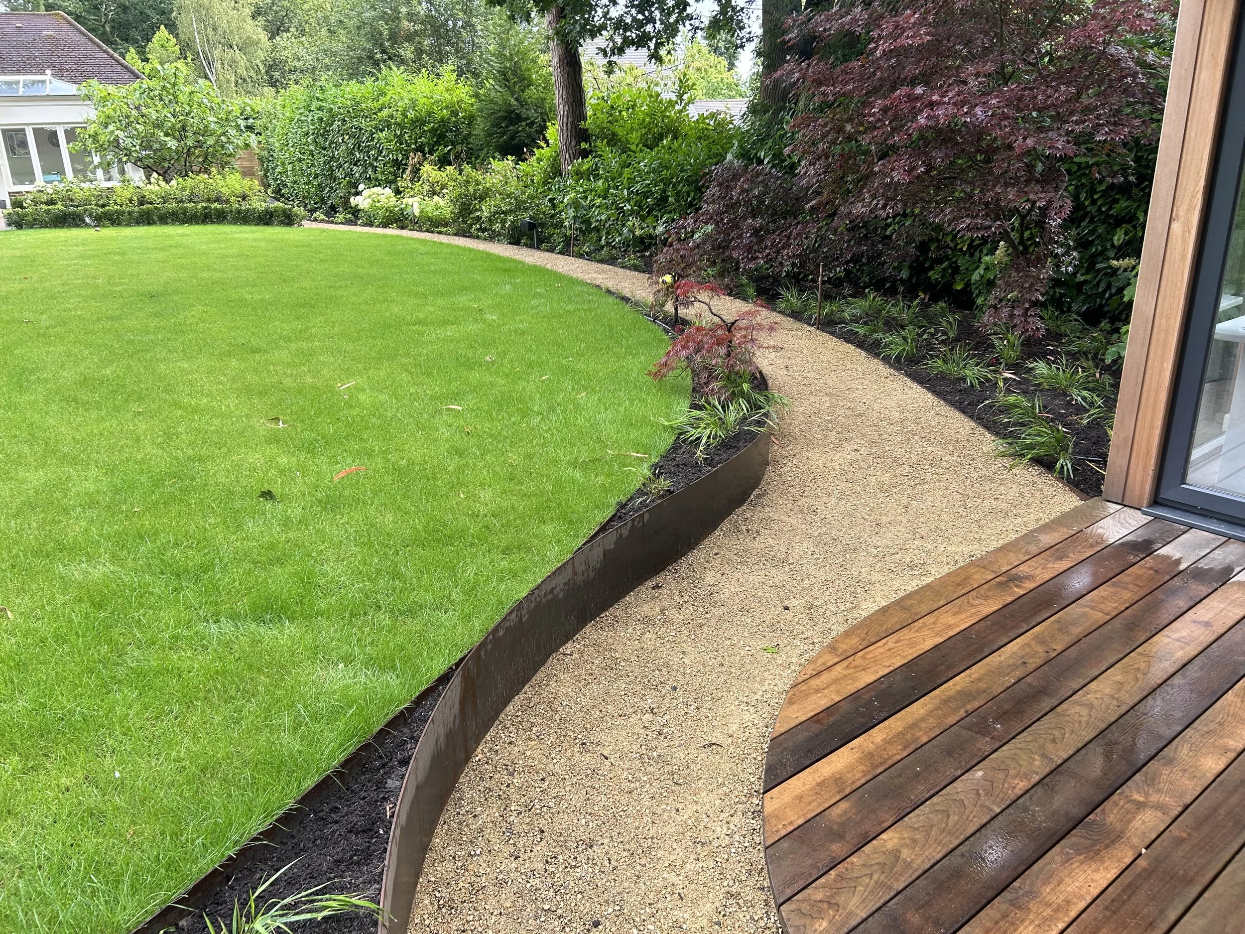A neatly maintained backyard with a curved garden bed, lush green grass, and a gravel pathway beside a wooden deck.