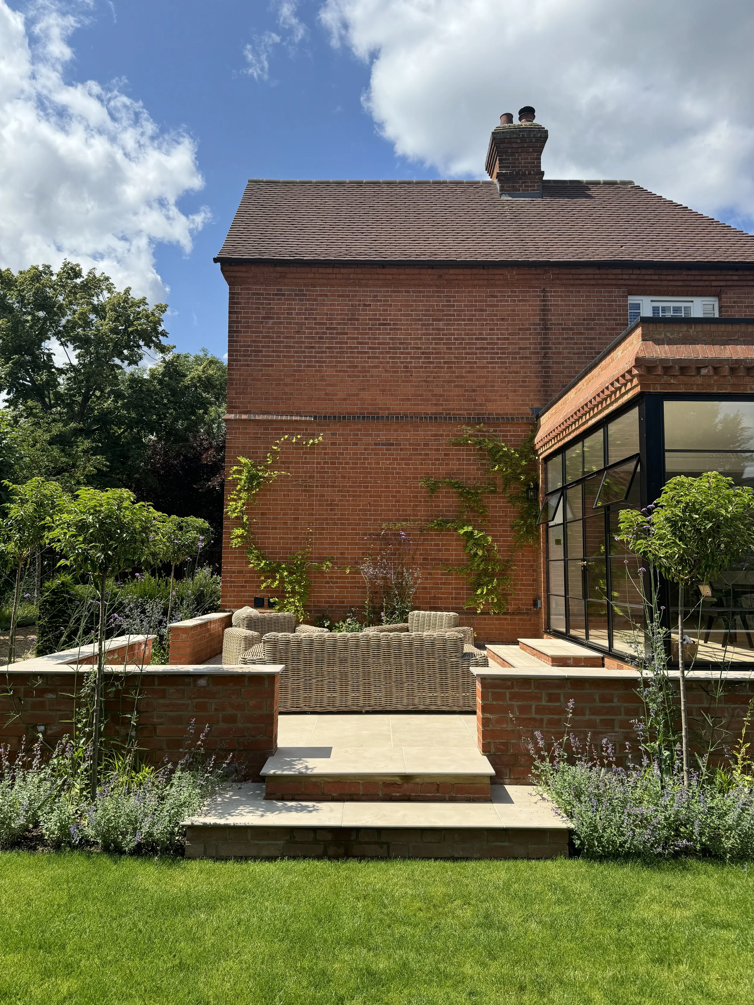 Backyard patio with wicker seating, brick steps, green plants, and a brick house under a partly cloudy sky.