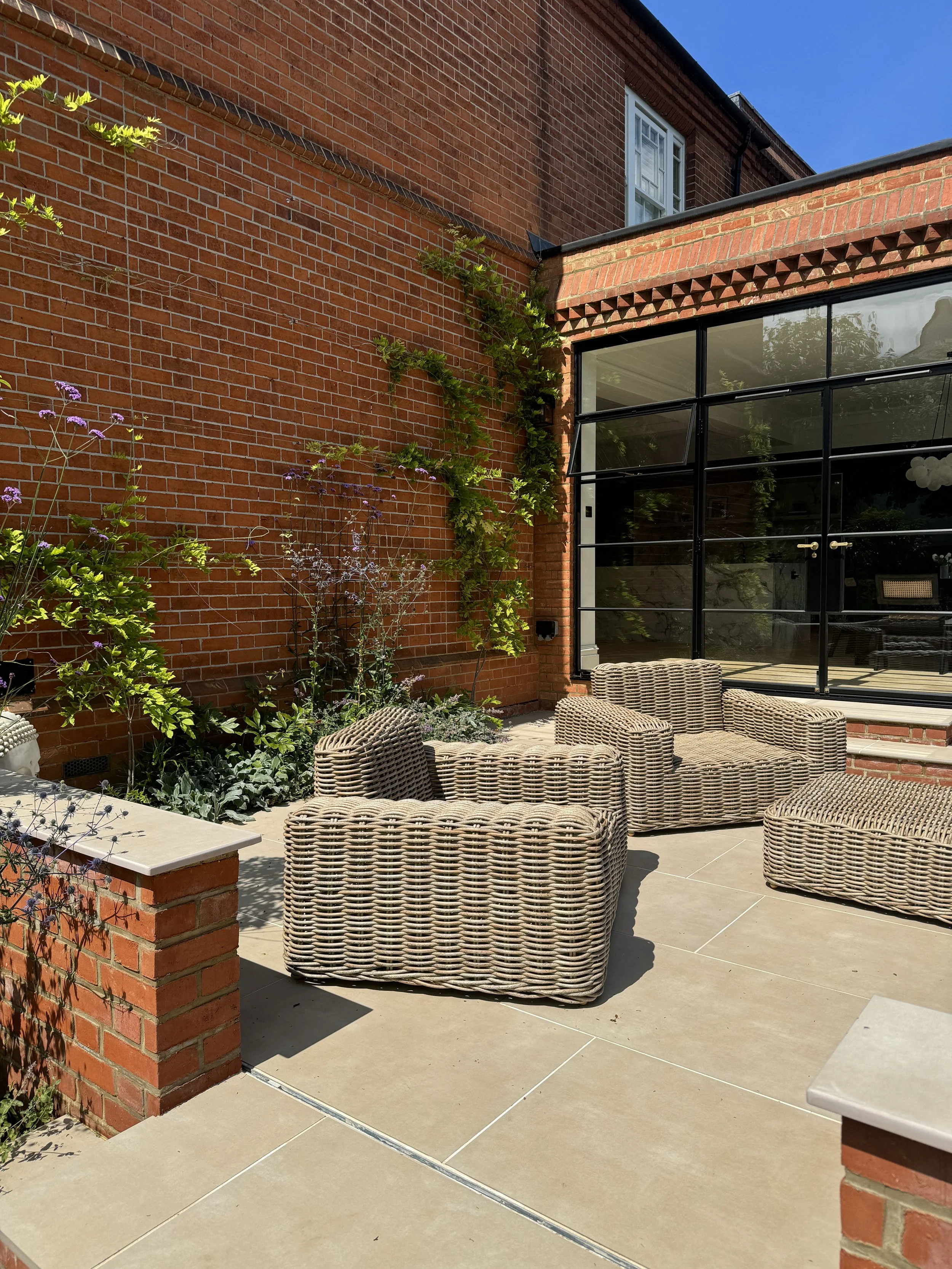 Outdoor patio with wicker chairs and a brick wall with climbing plants, large glass doors, and a bright blue sky.