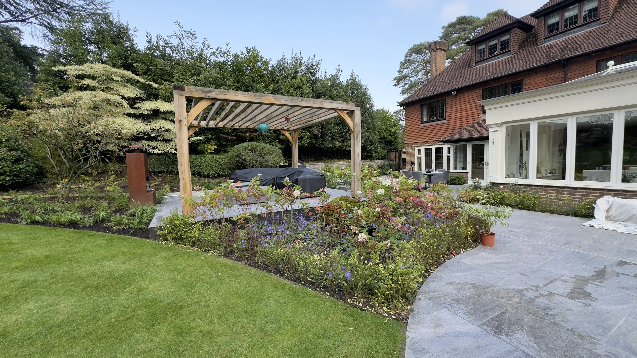 Backyard garden with a brick house, patio, wooden pergola, and flower bed with various plants and flowers.