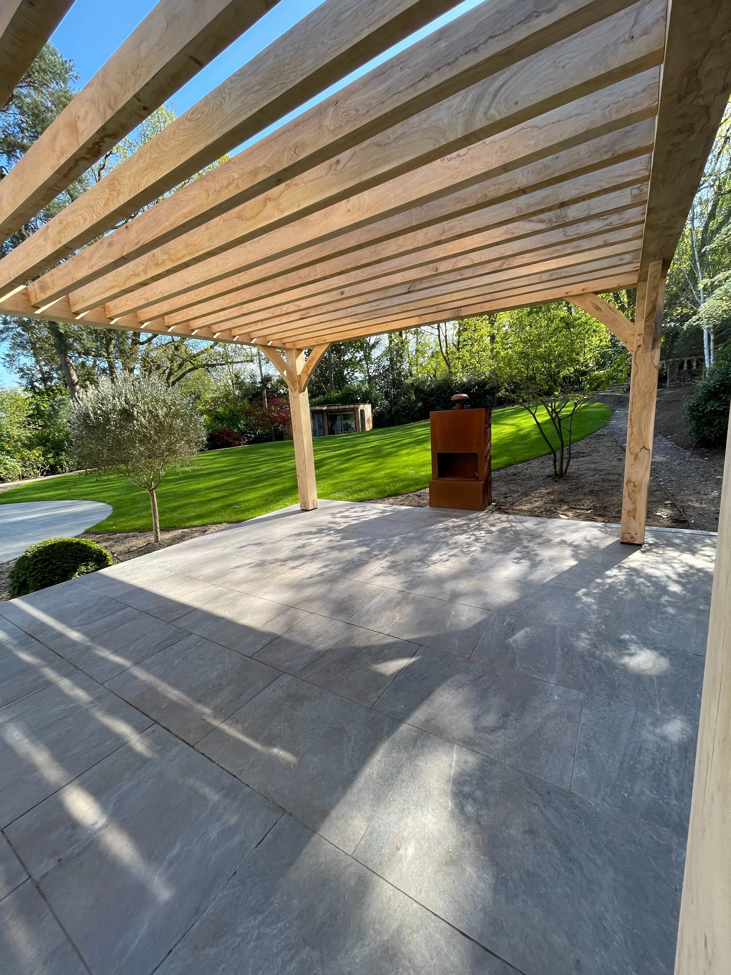 A wooden pergola with horizontal beams over a stone patio in a backyard with green grass, small trees, and shrubs under a clear blue sky.