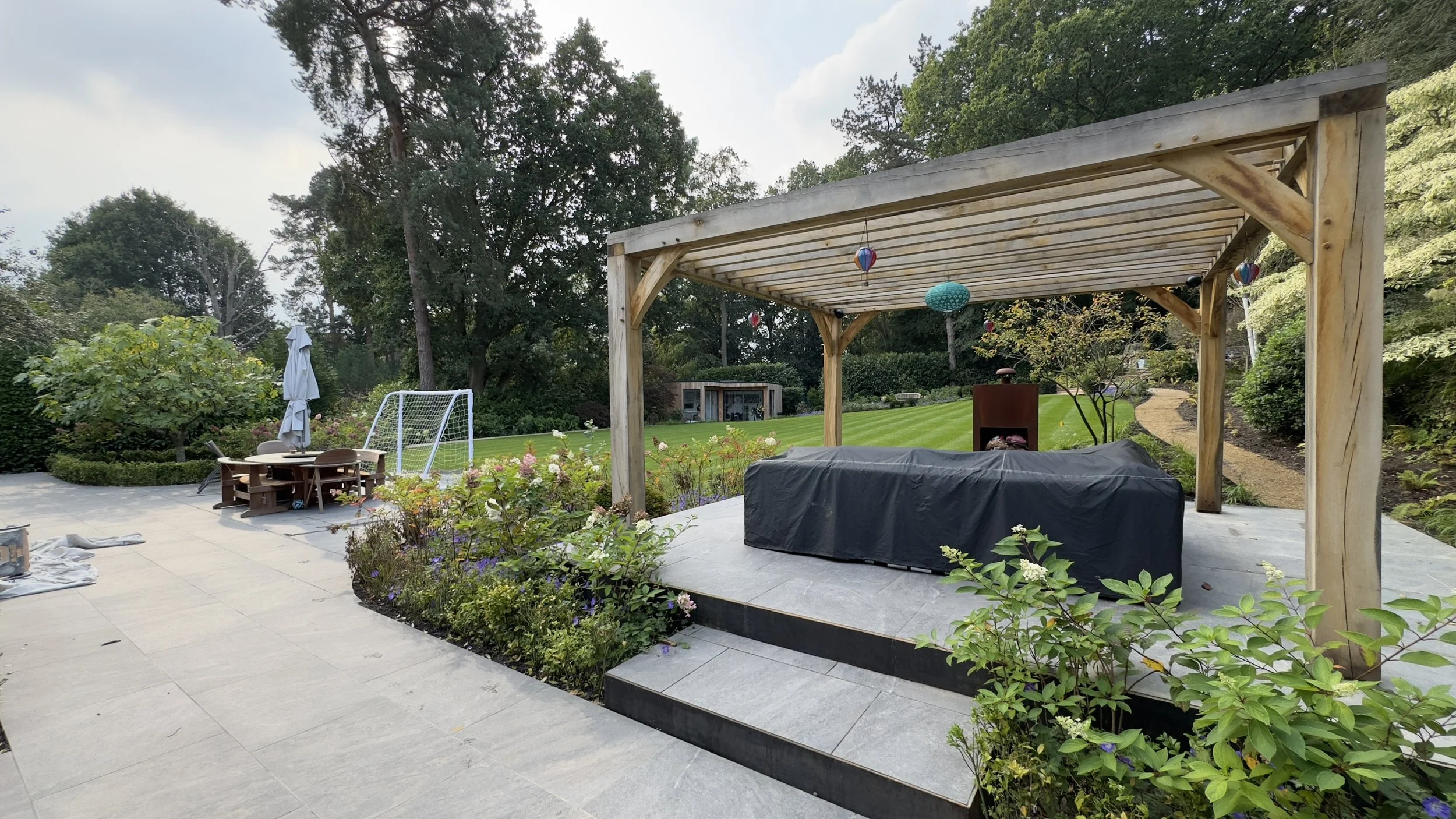 Backyard patio with a covered gazebo, outdoor furniture, a small soccer goal, and a lush green lawn with trees and shrubs in the background.