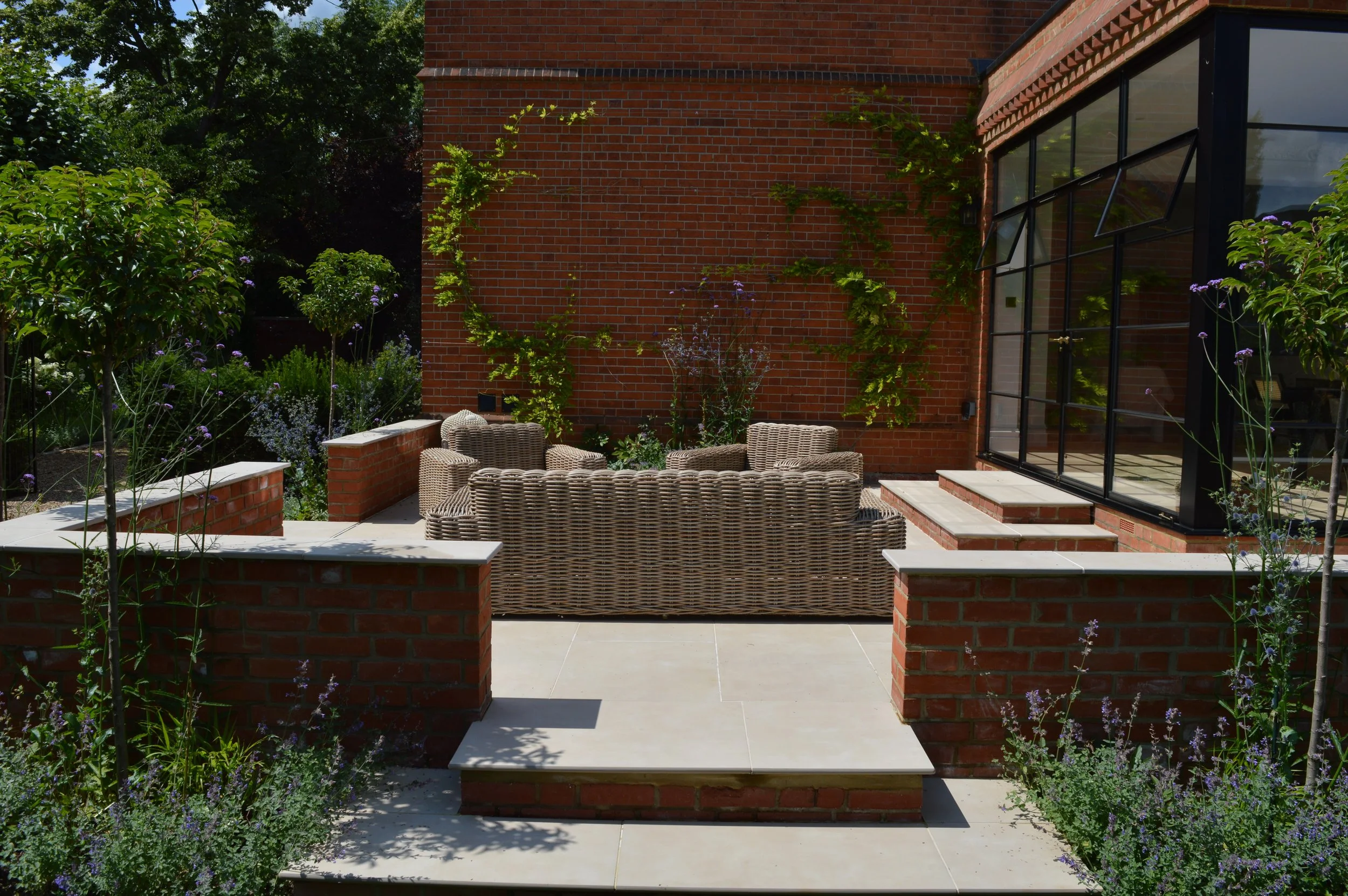 Outdoor patio area with wicker seating, surrounded by brick walls, stairs, potted plants, and flowering bushes in a garden setting.