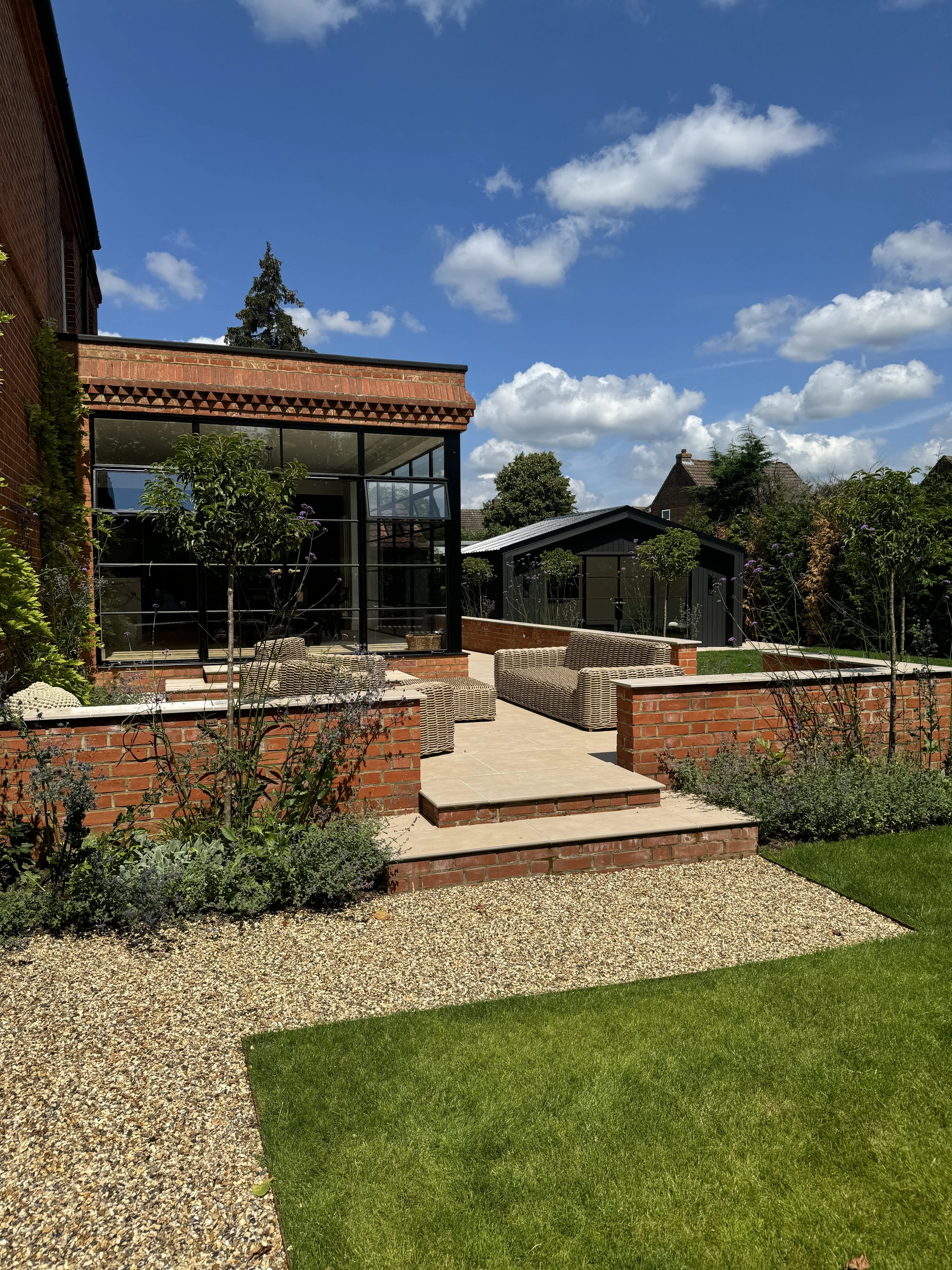 A backyard with a raised patio and tiered brick walls, wicker outdoor furniture, and a glass conservatory attached to a brick house, surrounded by plants and trees under a blue sky with scattered clouds.