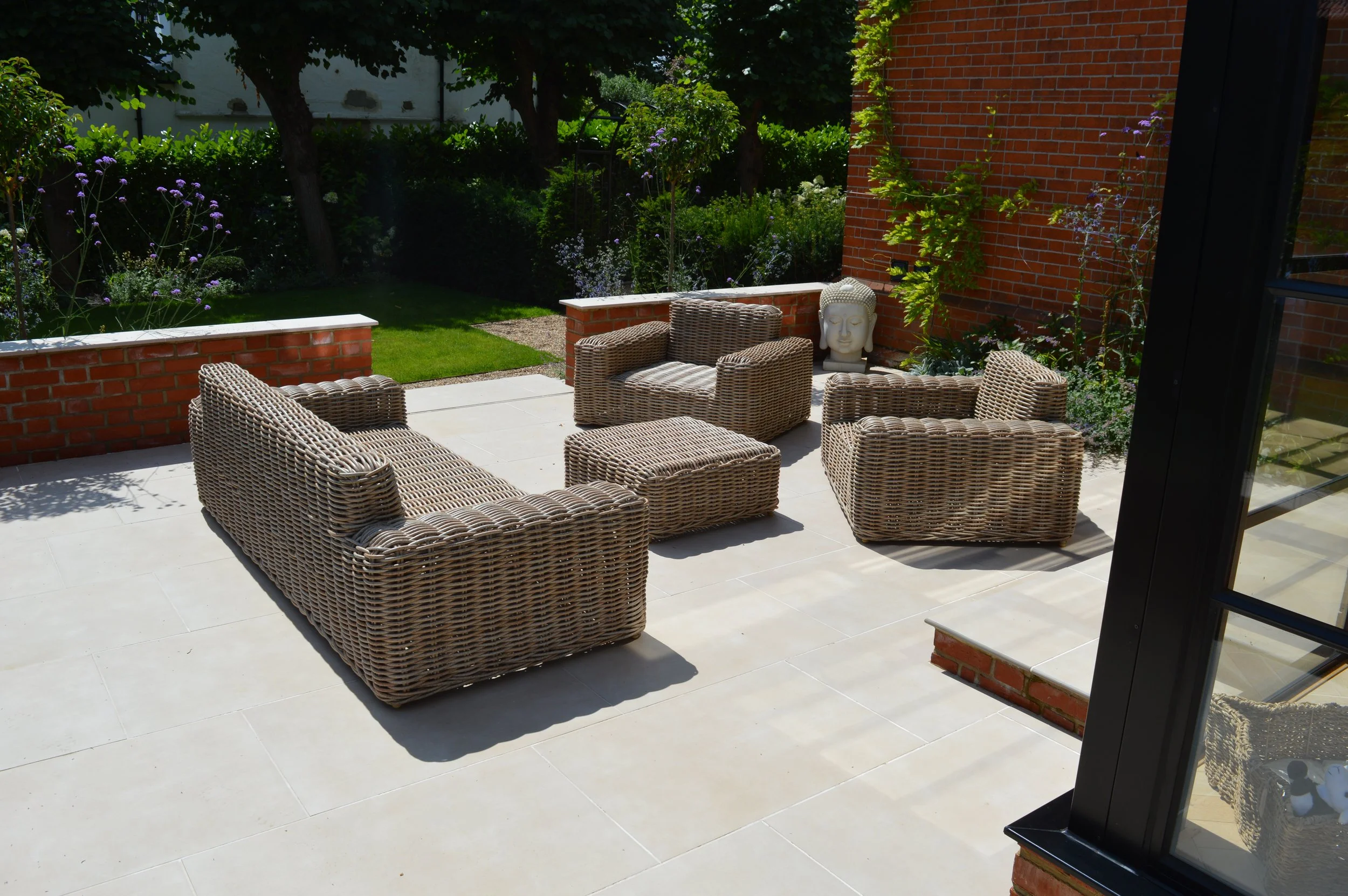 Outdoor patio with wicker furniture including a sofa, chairs, and ottomans, with a garden and trees in the background.