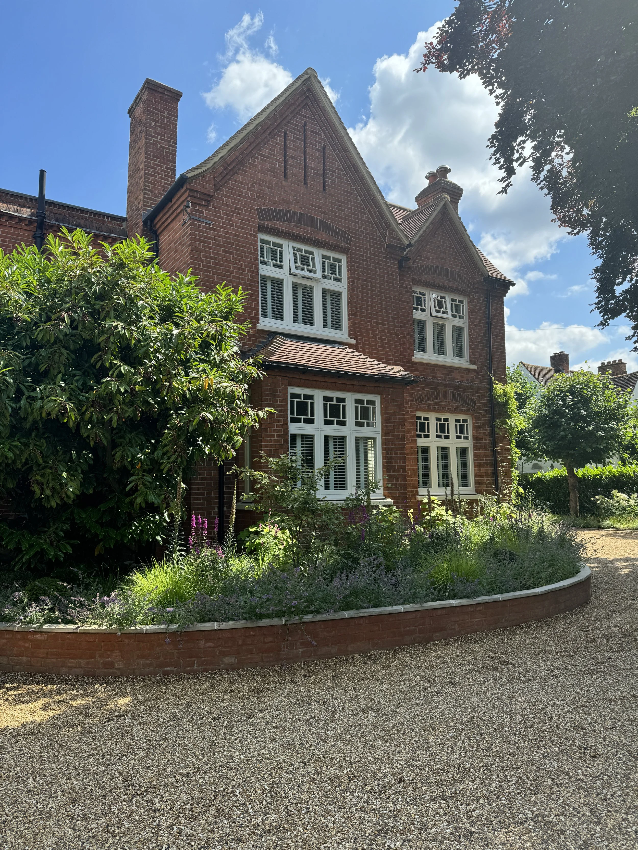 A two-story red brick house with white-framed windows, a garden with bushes and flowers, and a gravel driveway under a partly cloudy sky.