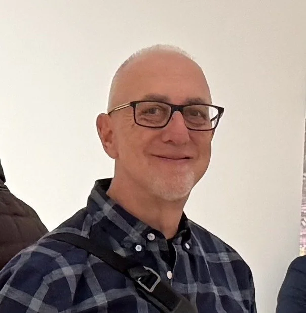 A middle-aged man wearing glasses and a checkered shirt, smiling indoors.