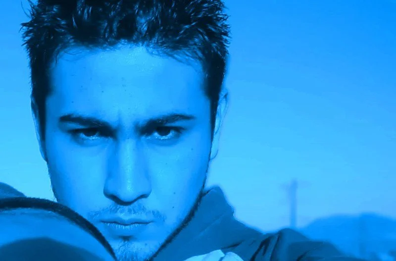 A young man with short dark hair and intense expression, looking directly into the camera, with a blue filter overlay.