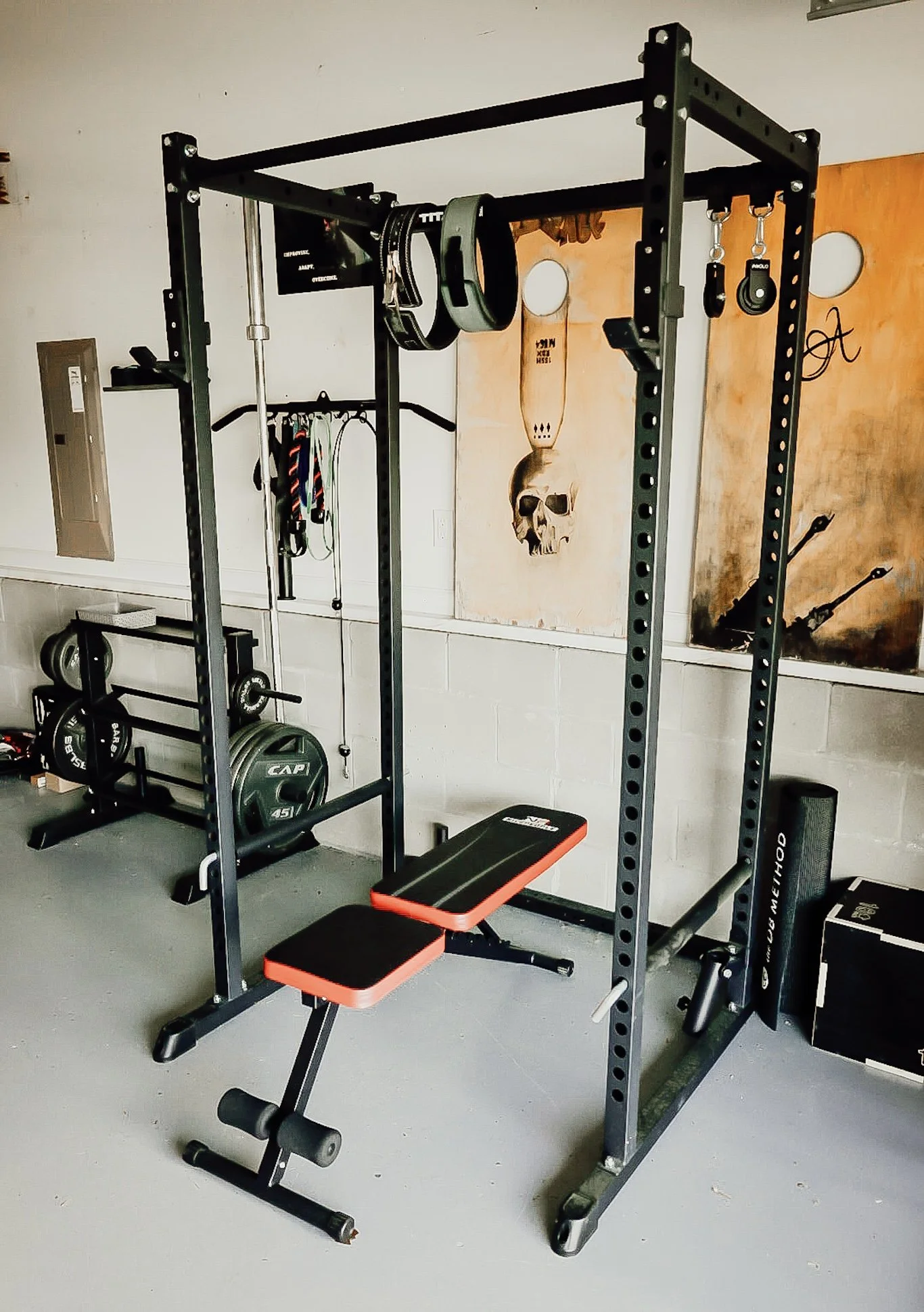 A home gym setup with a black power rack, weight plates, resistance bands, a bench with orange accents, and various workout equipment in a room with motivational posters and artwork on the wall.