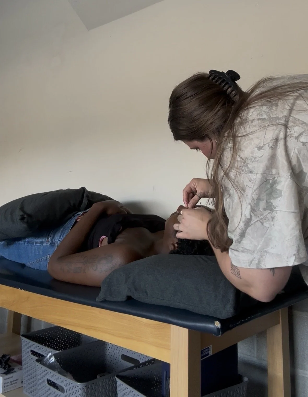 A woman with long hair and tattoos on her arm is examining or checking the mouth of a man lying on a bed or table, in a room with a beige wall.