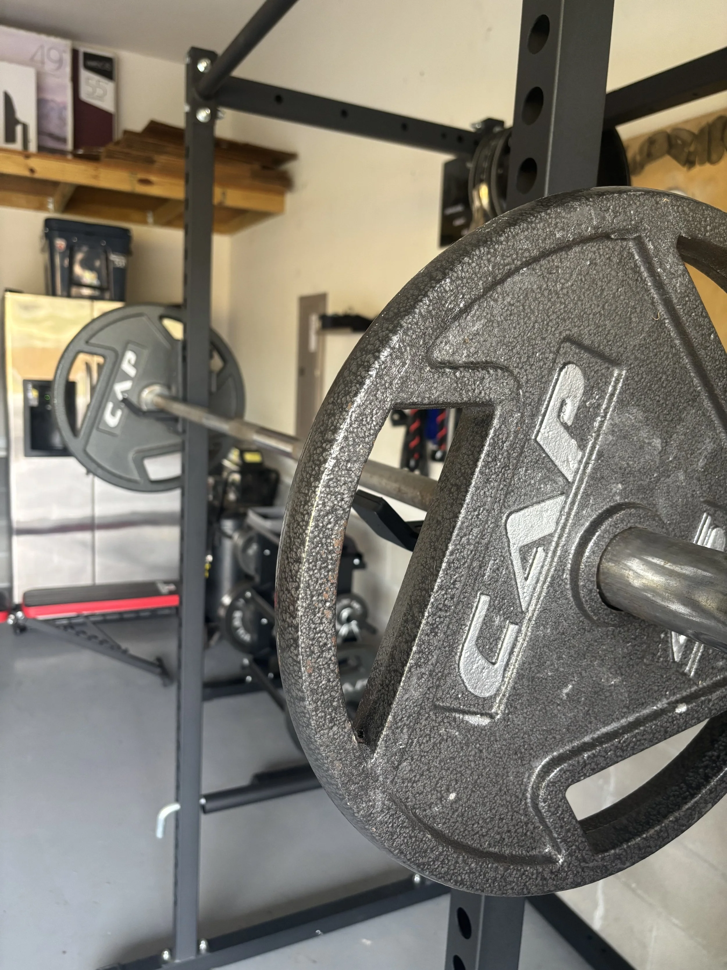 Close-up of a weightlifting barbell with a cast-iron weight on it, in a garage gym with a weight rack and storage shelves in the background.