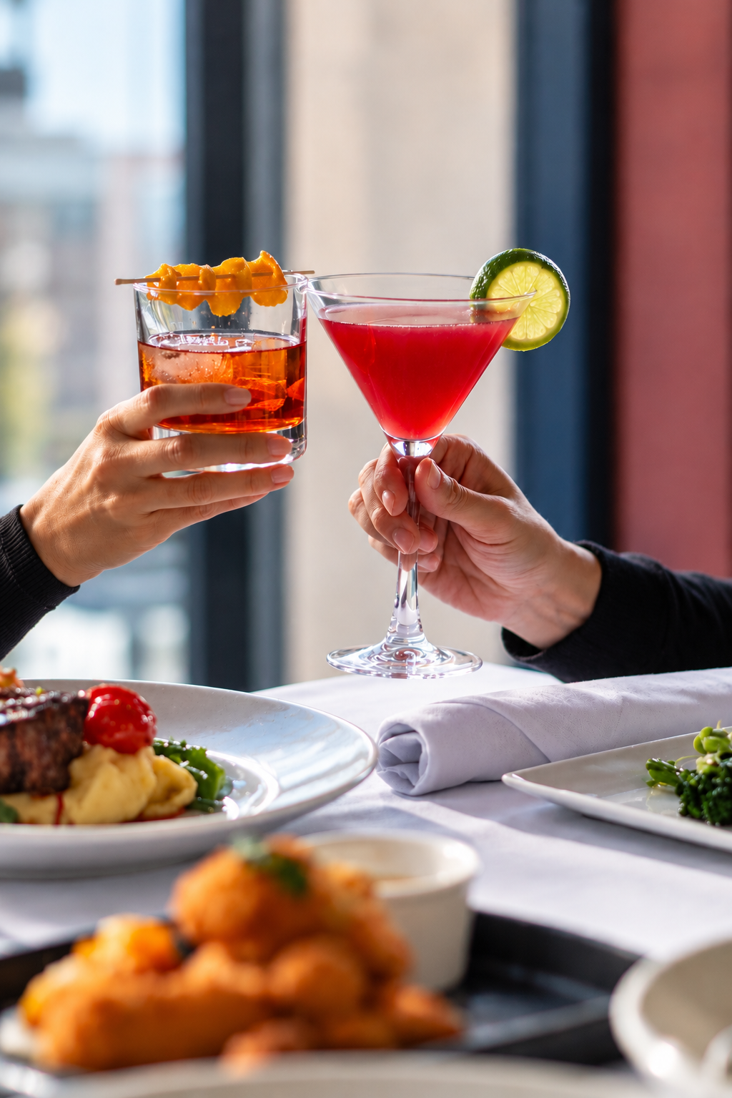 People toasting with cocktails, one in a martini glass garnished with a lime wheel and the other in a glass with an orange peel garnish.