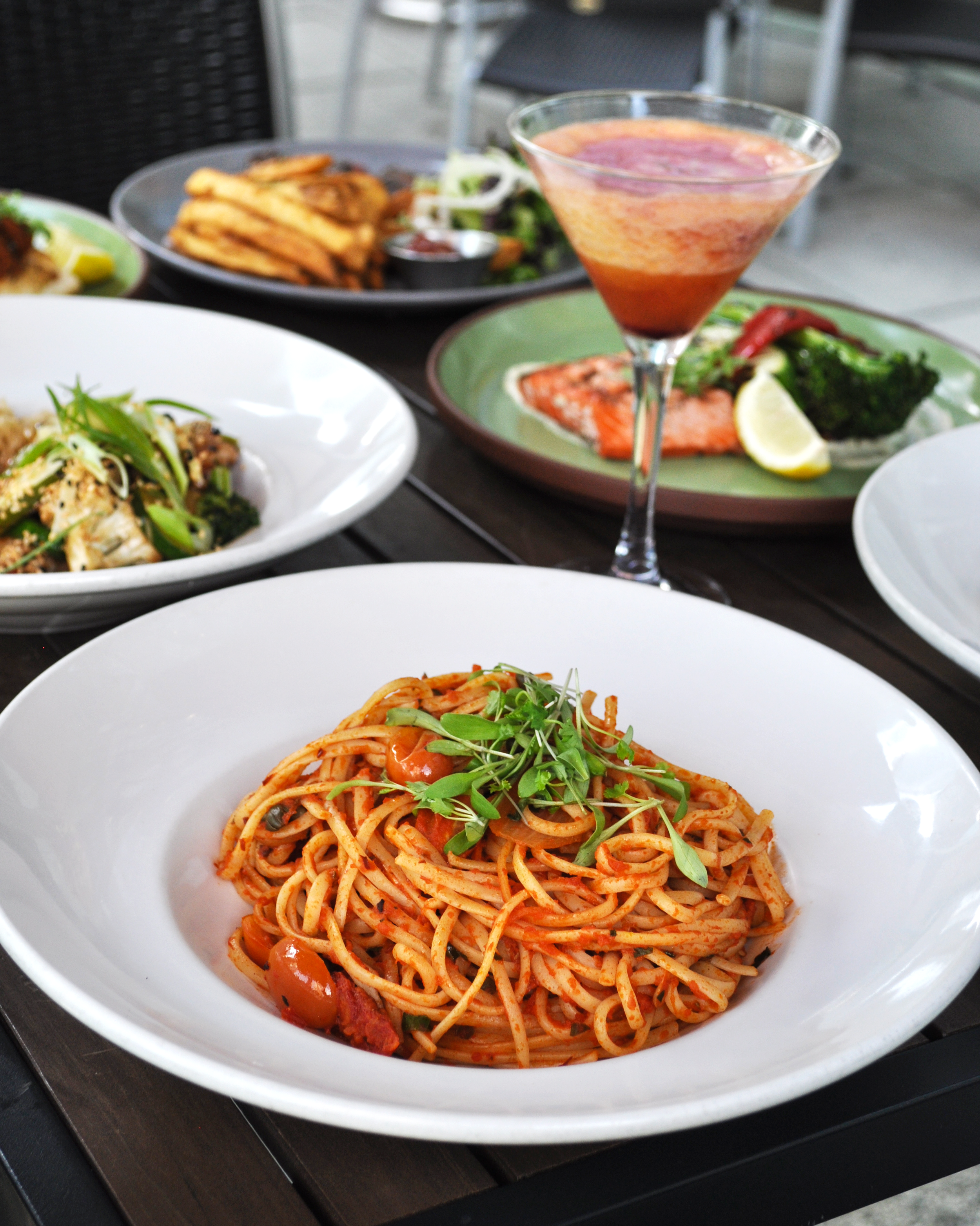 A delicious selection of freshly prepared dishes served on a welcoming outdoor restaurant patio. A vibrant bowl of pasta tossed in a rich tomato sauce, topped with fresh herbs and cherry tomatoes, takes centre stage, surrounded by beautifully plated 