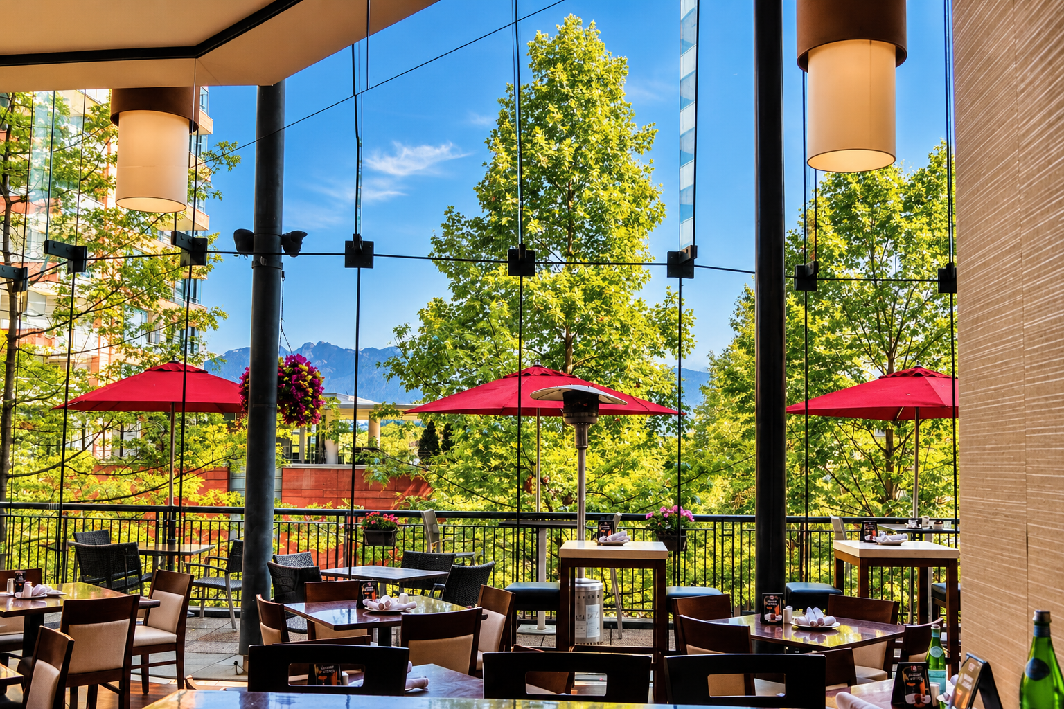 A bright, inviting restaurant and patio space featuring floor-to-ceiling glass windows, modern furnishings, and vibrant red patio umbrellas. Surrounded by lush greenery with scenic mountain views in the distance, the space offers a welcoming atmosphe