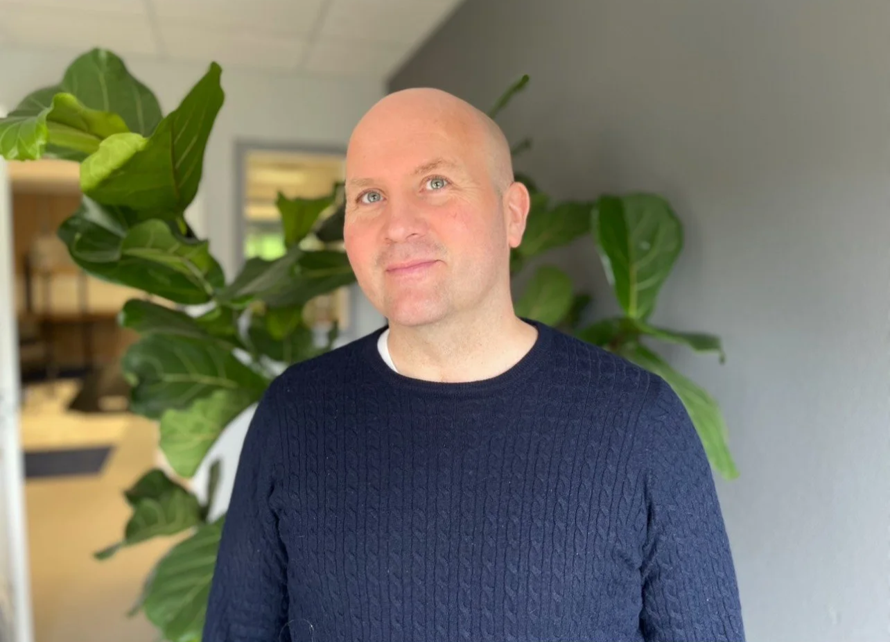 En mann med lyst hår og blå øyne, iført en mørk blå genser, står foran grønne planter i et rom.