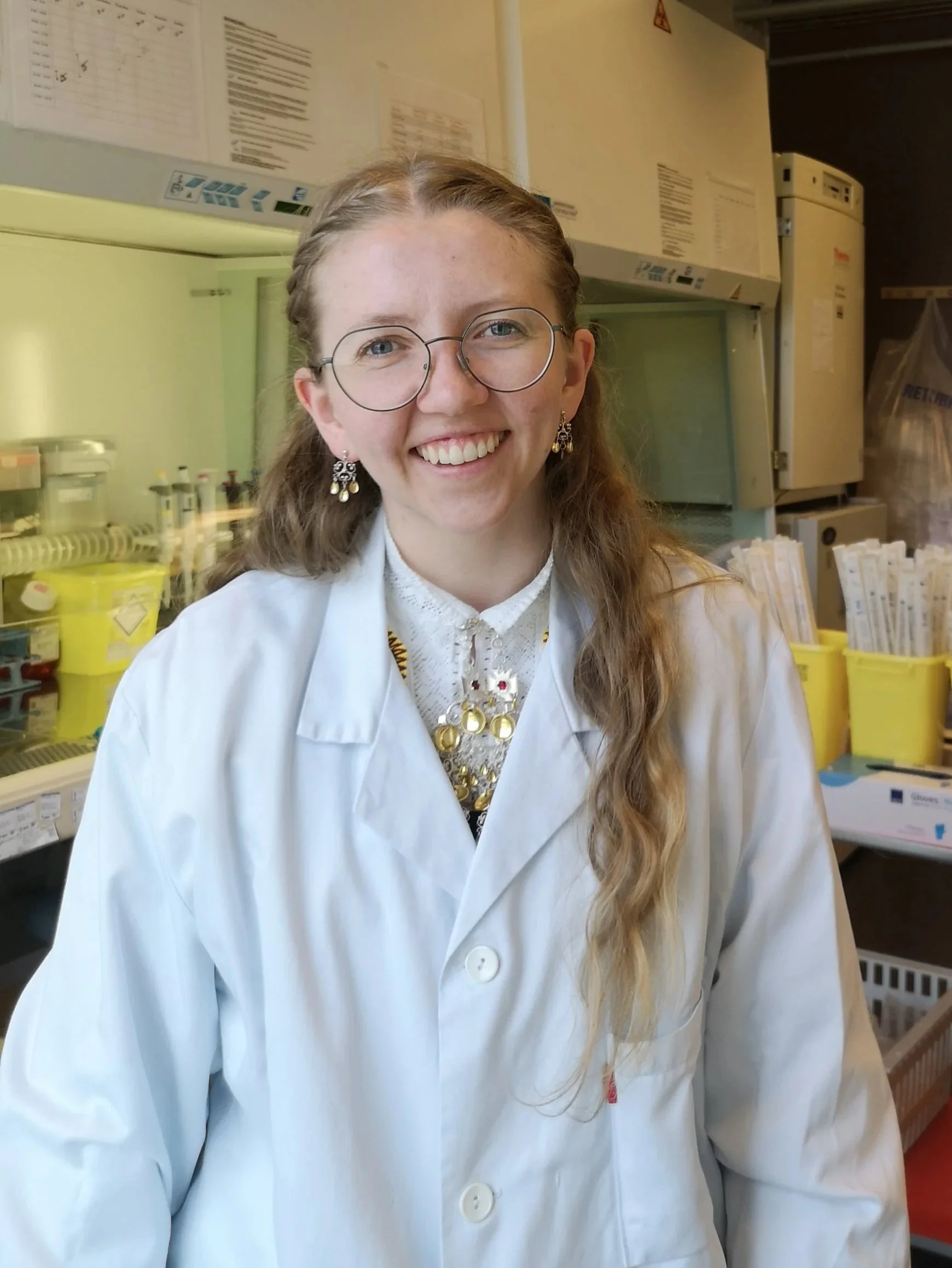 En kvinne med brunt, krøllete hår, iført hvit labfrakk og store rundt briller poserer på et laboratorium med laboratorieutstyr og hyller i bakgrunnen.