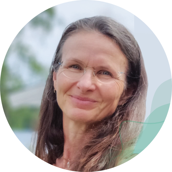 Et portrett av en smilende kvinne med brunt hår og briller, utendørs med grønn natur i bakgrunnen.