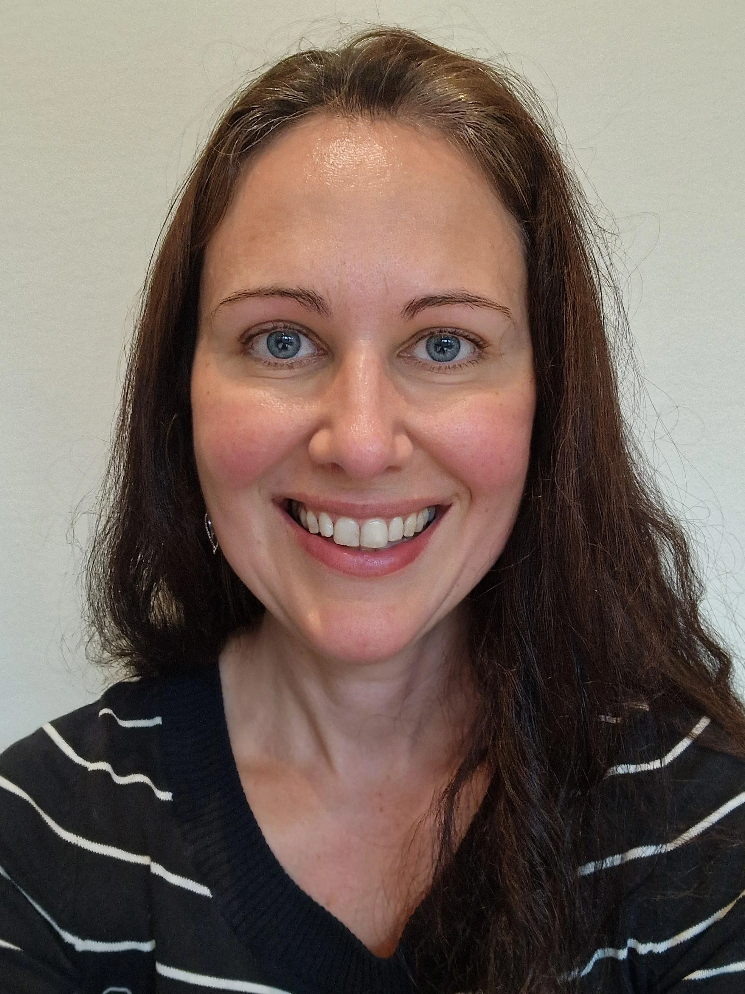 Et portrett av en smilende kvinne med brunt, langt, bølget hår, en svart og hvit stripete genser, og blå øyne, fotografert foran en lys vegg.