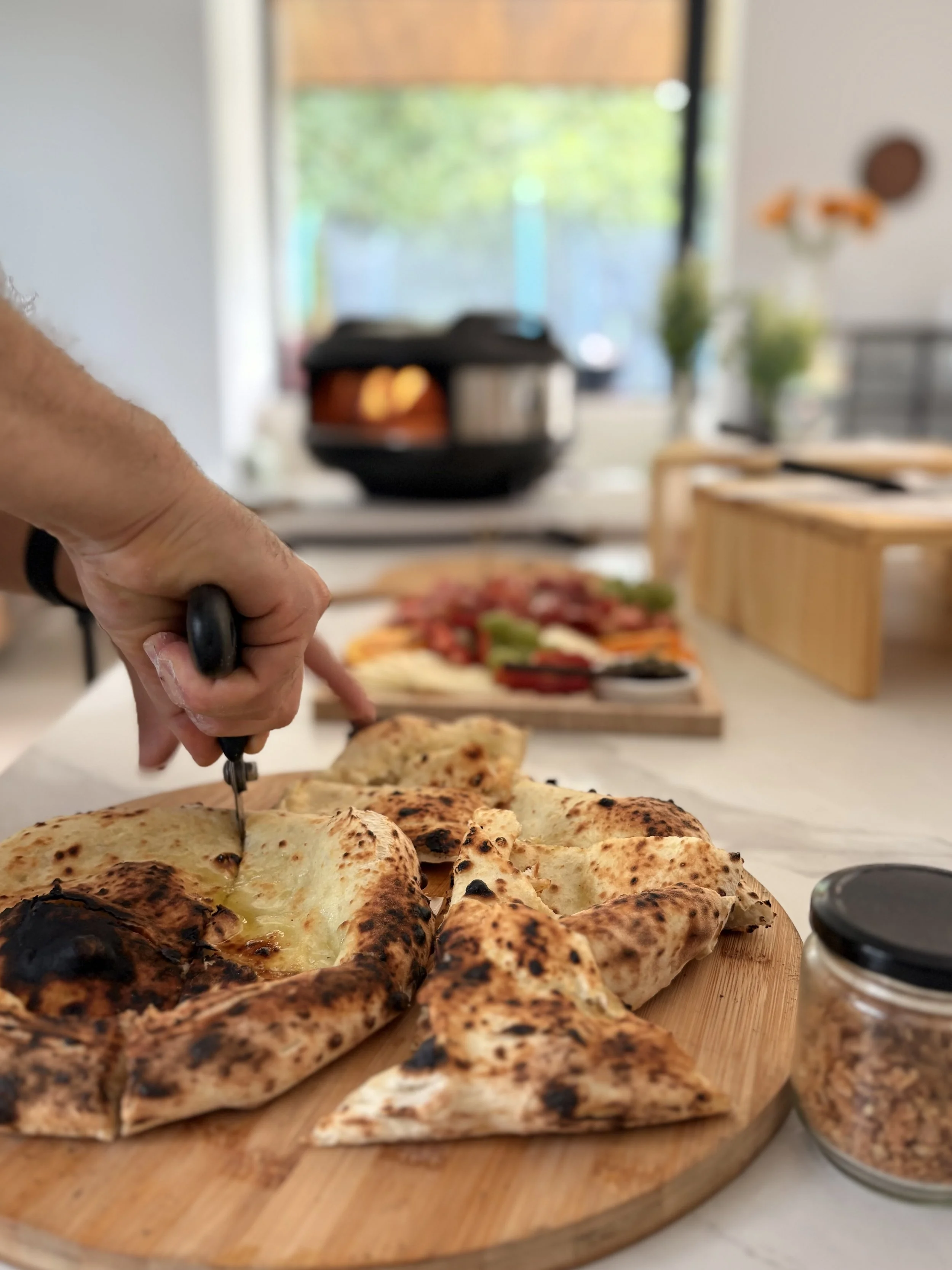 Persona cortando una pizza en una tabla de madera en una cocina iluminada naturalmente, con ingredientes y utensilios en el fondo.