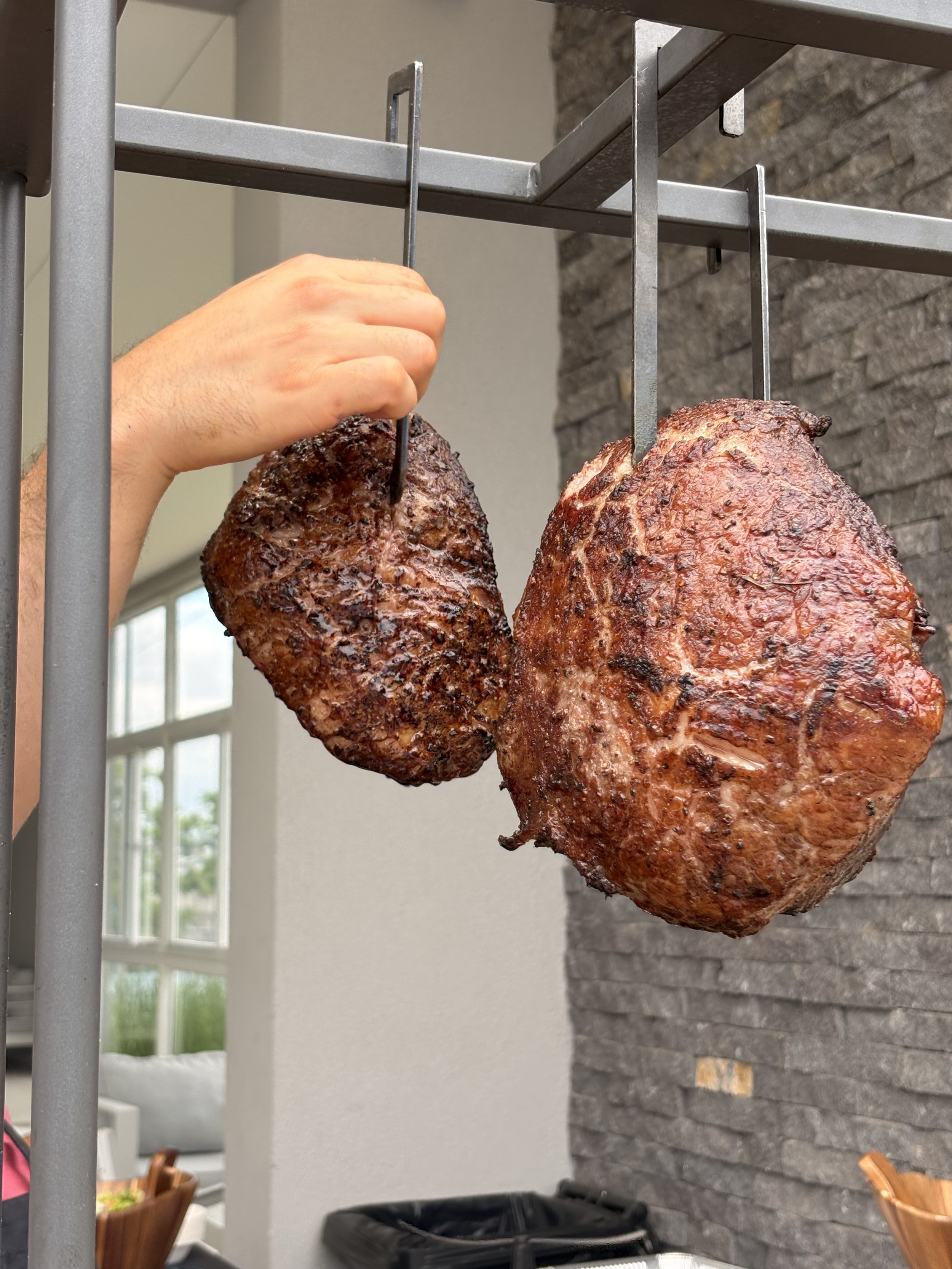 Dos piezas grandes de carne asada colgadas en un asador de barbacoa, con una mano ajustando una de ellas.
