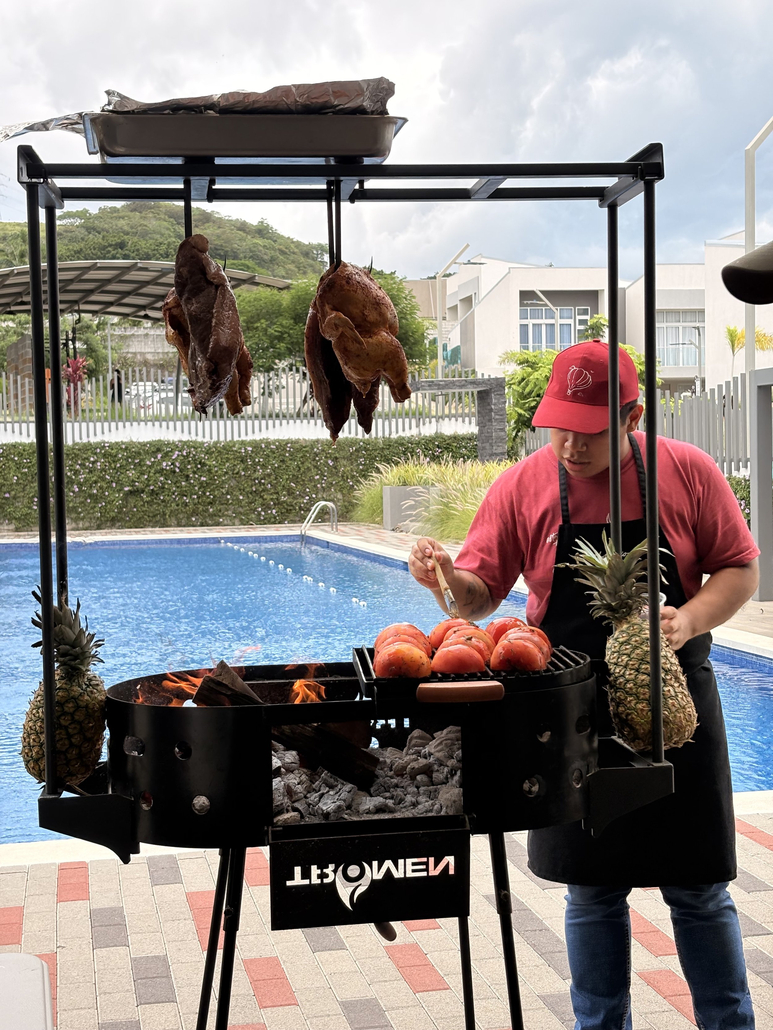 Persona sirviendo comida en un asador con carnes colgadas y frutas, con piscina y casas en el fondo en un día nublado.