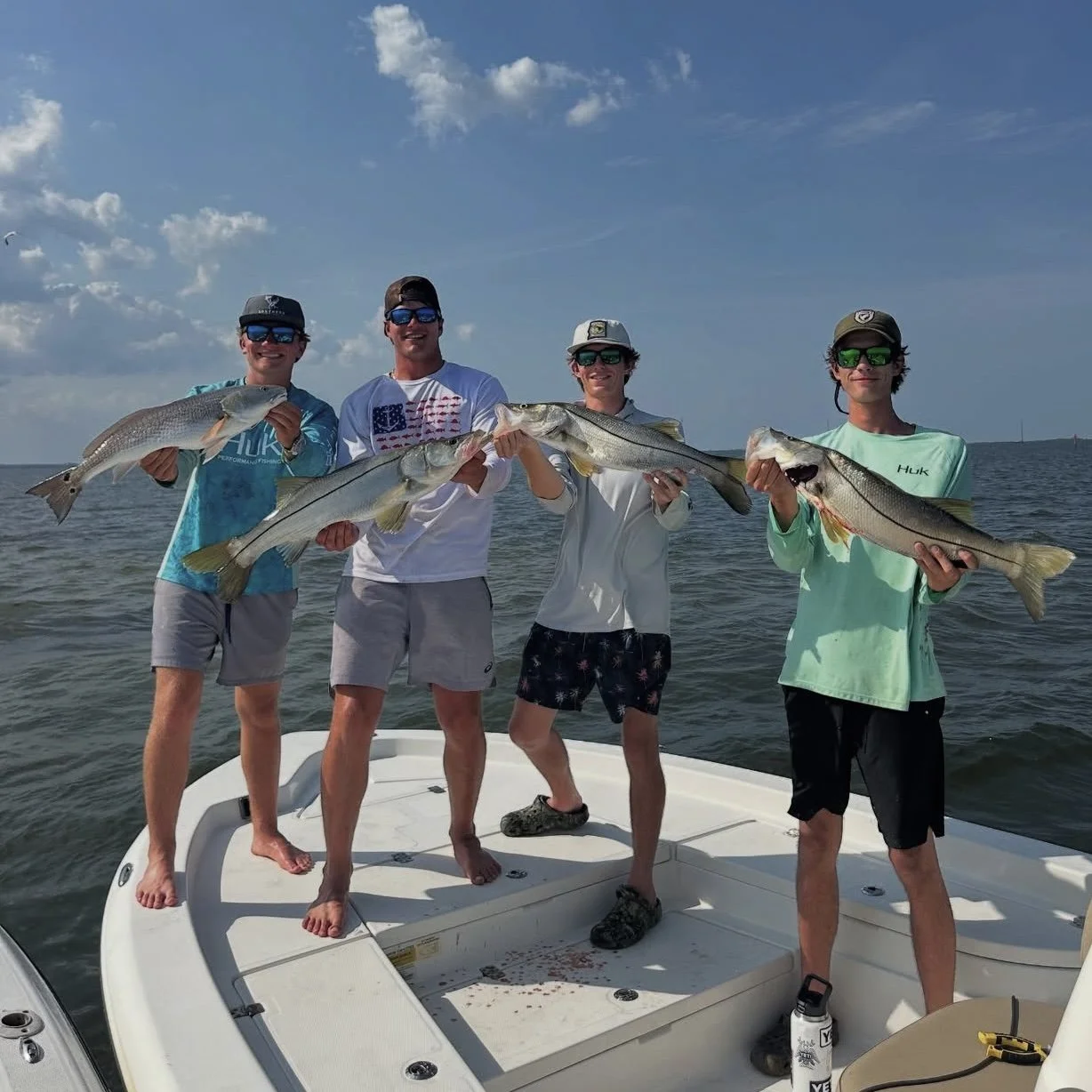 Four men on a boat holding large fish they caught, under a partly cloudy sky.