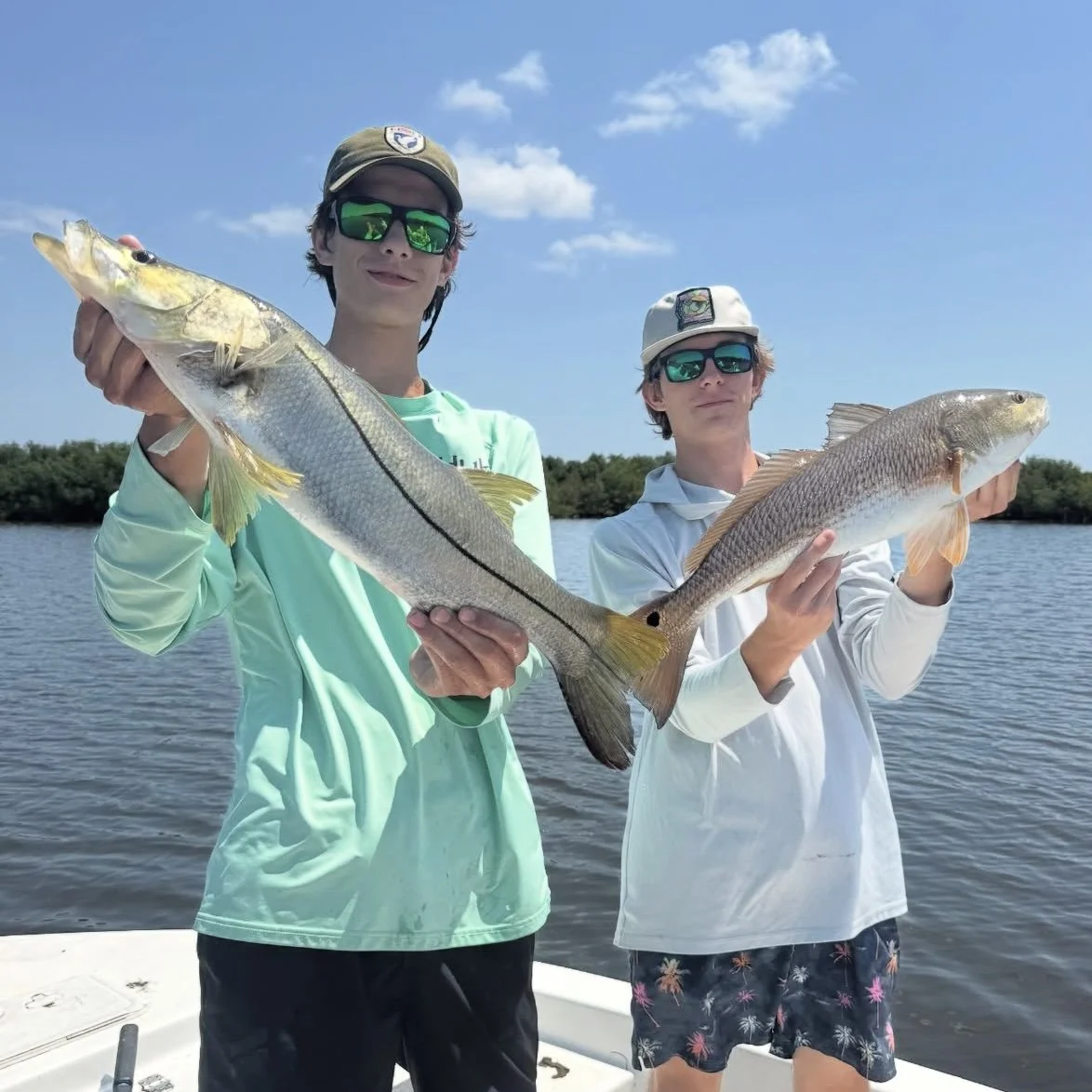 Two young men on a boat holding up large fish they caught, with water and trees in the background under a blue sky with clouds.