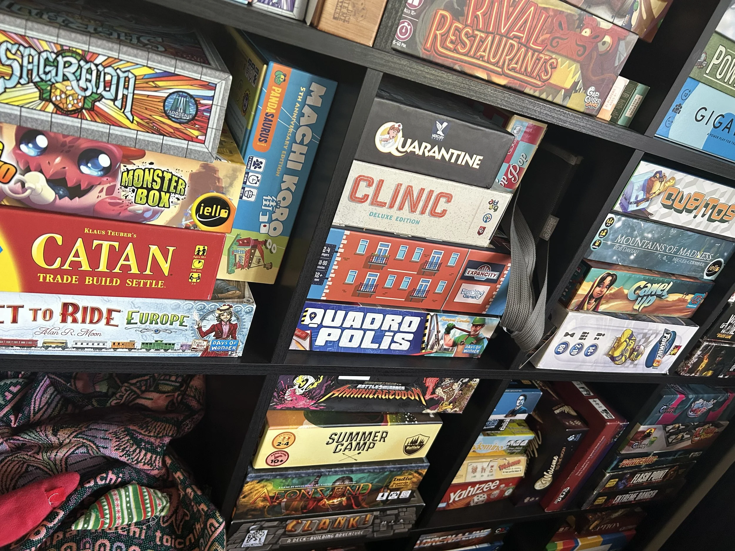 Just like a Black Meeple, I have over one hundred board games. This picture is a small portion of my collection. A few of the games featured are Clinic, Machi Koro, Quadropolis, Catan, Sagrada, Summer Camp, Aeons End, Clank, and Expancity.