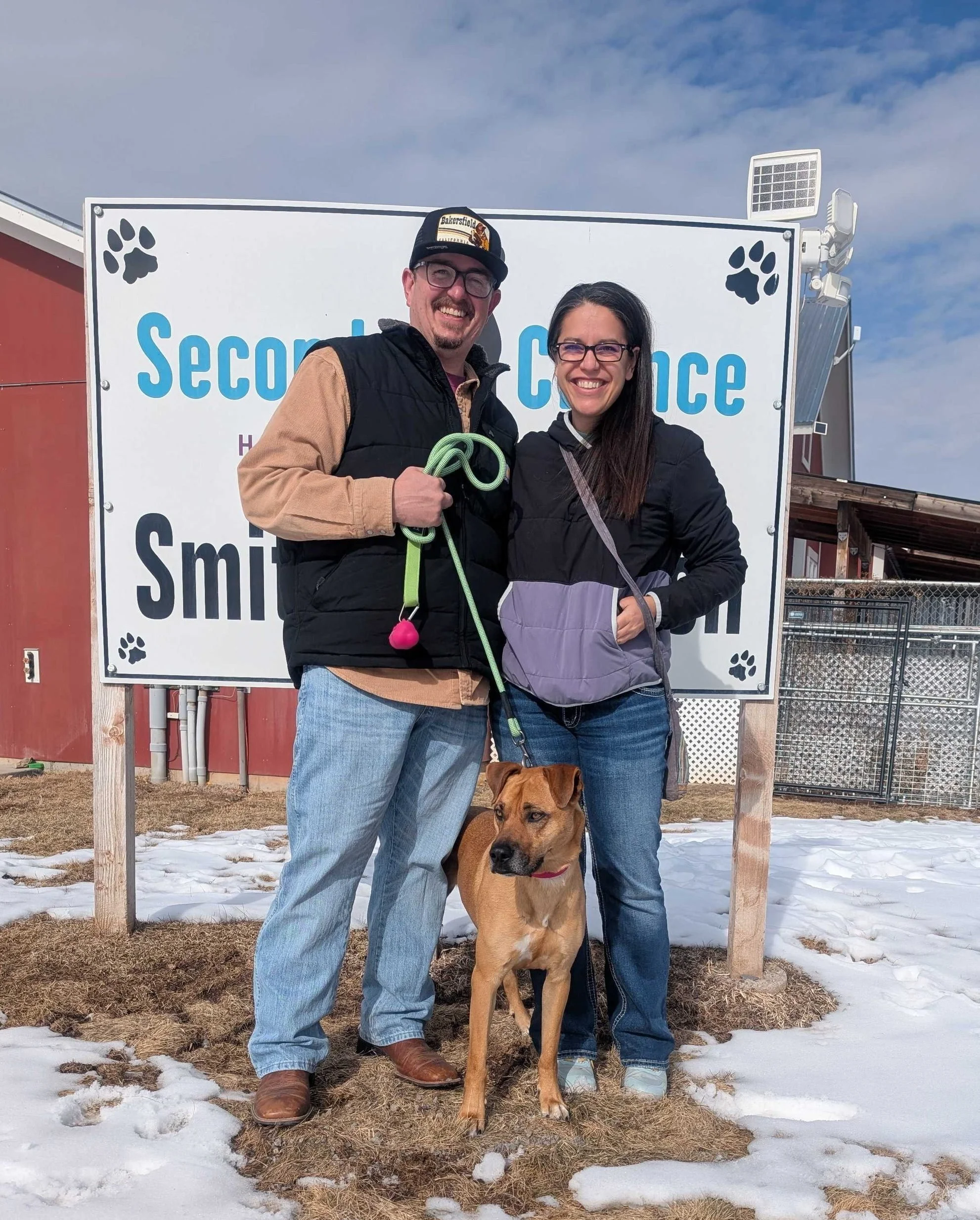 A man and a woman standing outside in front of Second Chance Humane Society sign with a tan dog on a leash. They are smiling in front of a sign that reads 'Second Chance Shelter' with paw print icons.