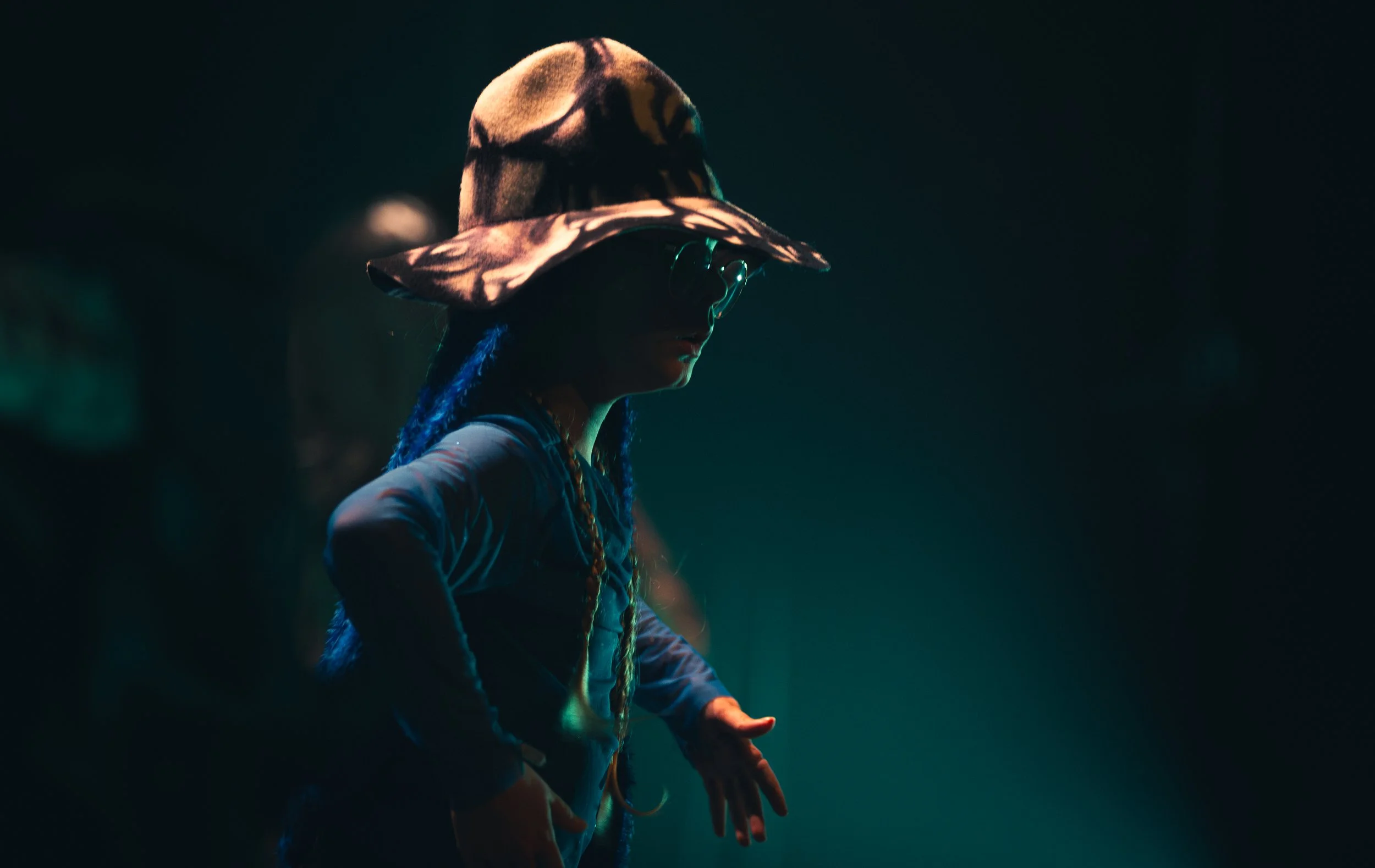 Personne portant un chapeau ample avec un motif camouflage ou abstrait, se trouvant dans un environnement sombre avec une lumière tamisée