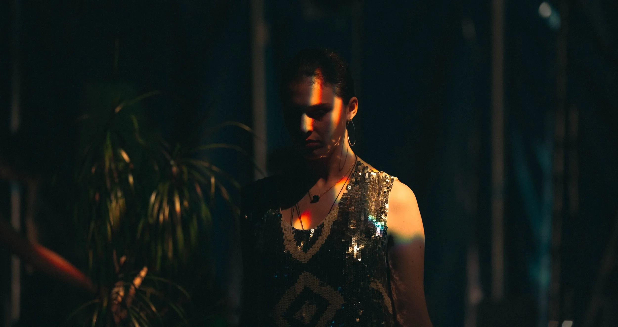 Une femme portant une robe à sequins noires et or, dans un environnement sombre avec un éclairage chaud, à côté d'une plante verte