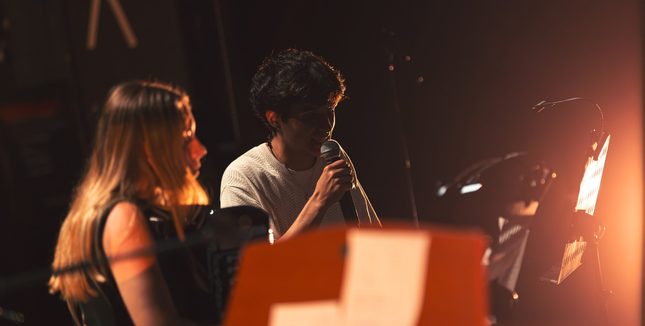 Deux personnes, une femme blonde et une homme aux cheveux foncés, discutent au microphone dans un studio d'enregistrement, avec des partitions de musique sur un pupitre.