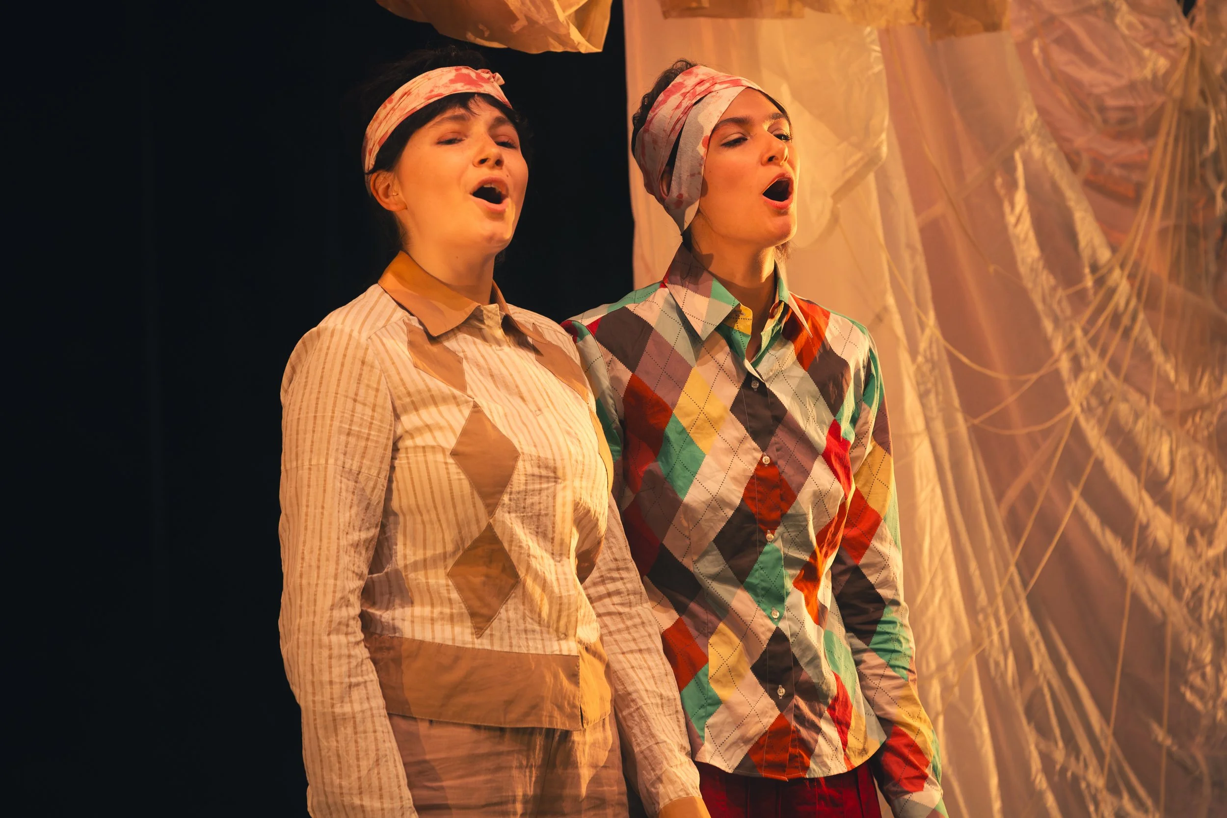 Deux femmes en chemises colorées chantent sur scène lors d'une performance, avec un décor de tissu il y a derrière elles.