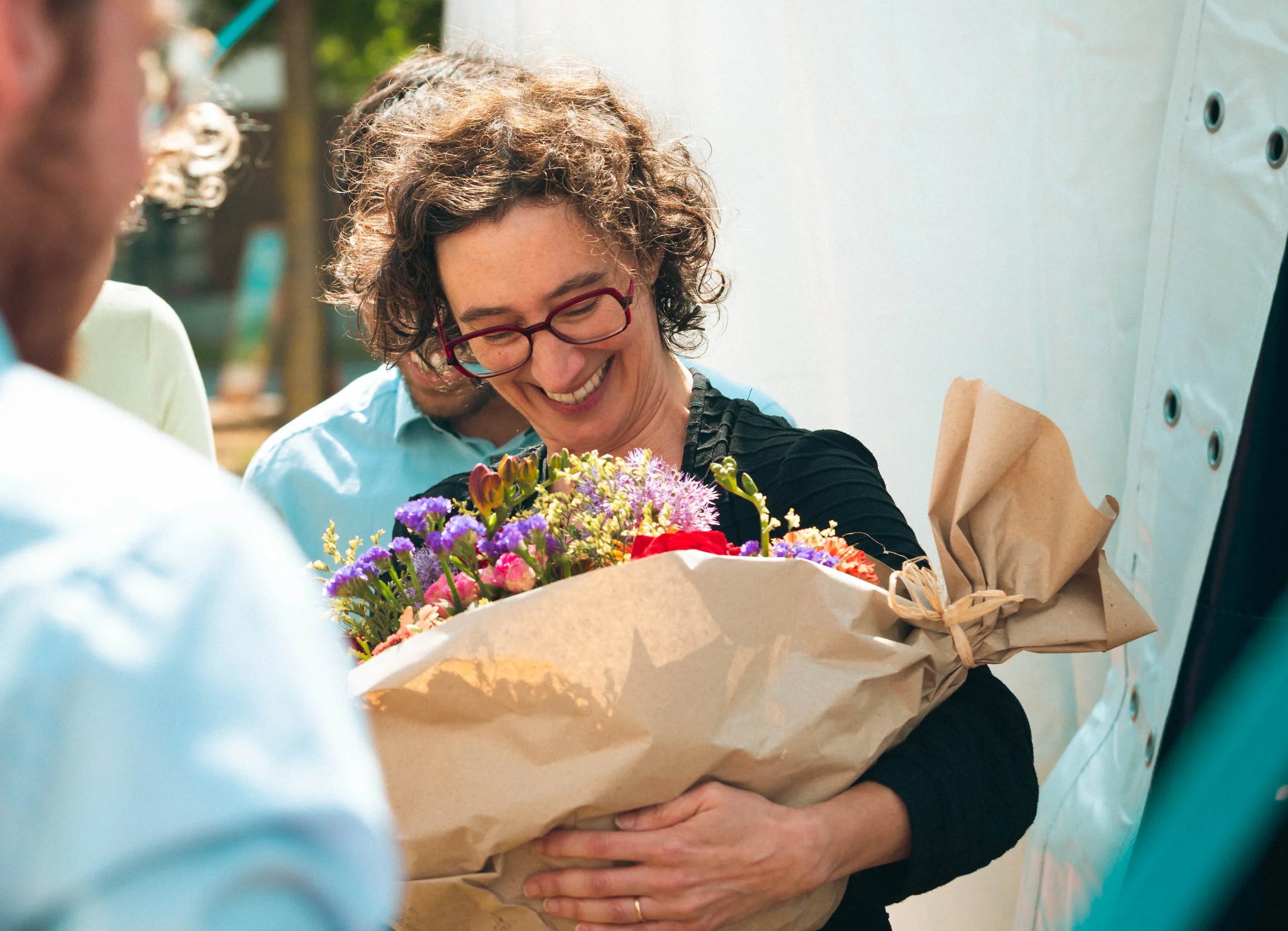 Une femme souriante tient un bouquet de fleurs dans ses bras lors d'une célébration extérieure.