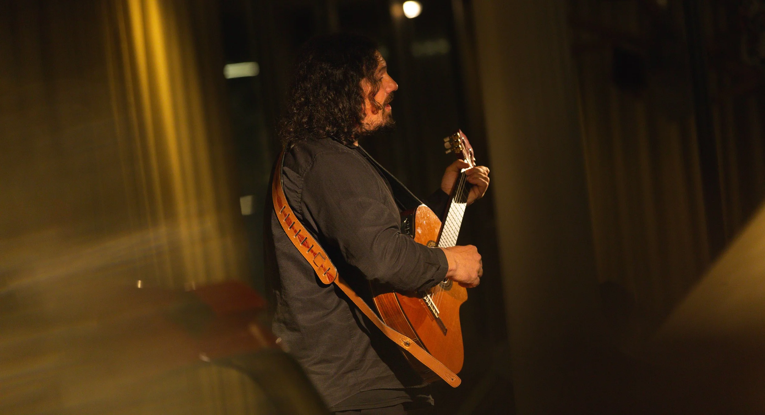 Un homme joue de la guitare acoustique dans une ambiance sombre, éclairé par une lumière chaude, avec des arrière-plans flous.