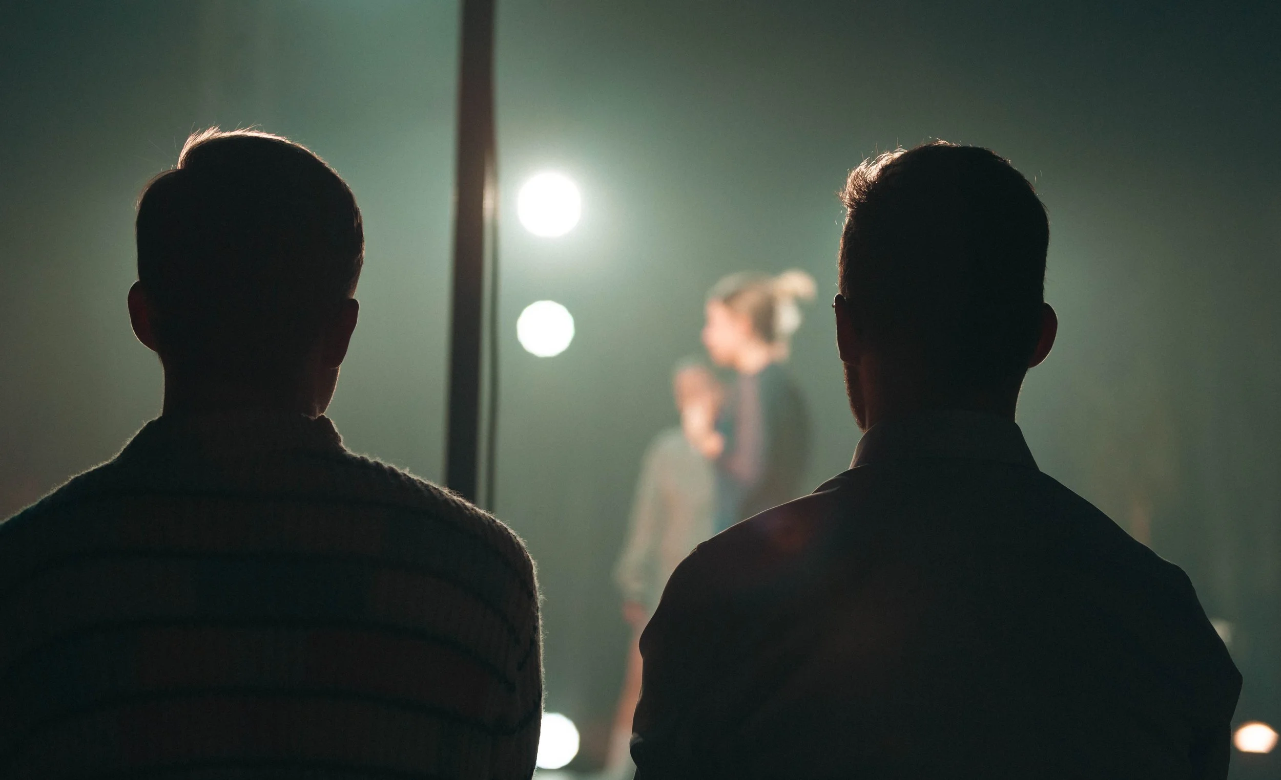 Deux hommes regardant un spectacle de scène en arrière-plan, une femme en costume de scène performe sous des lumières.