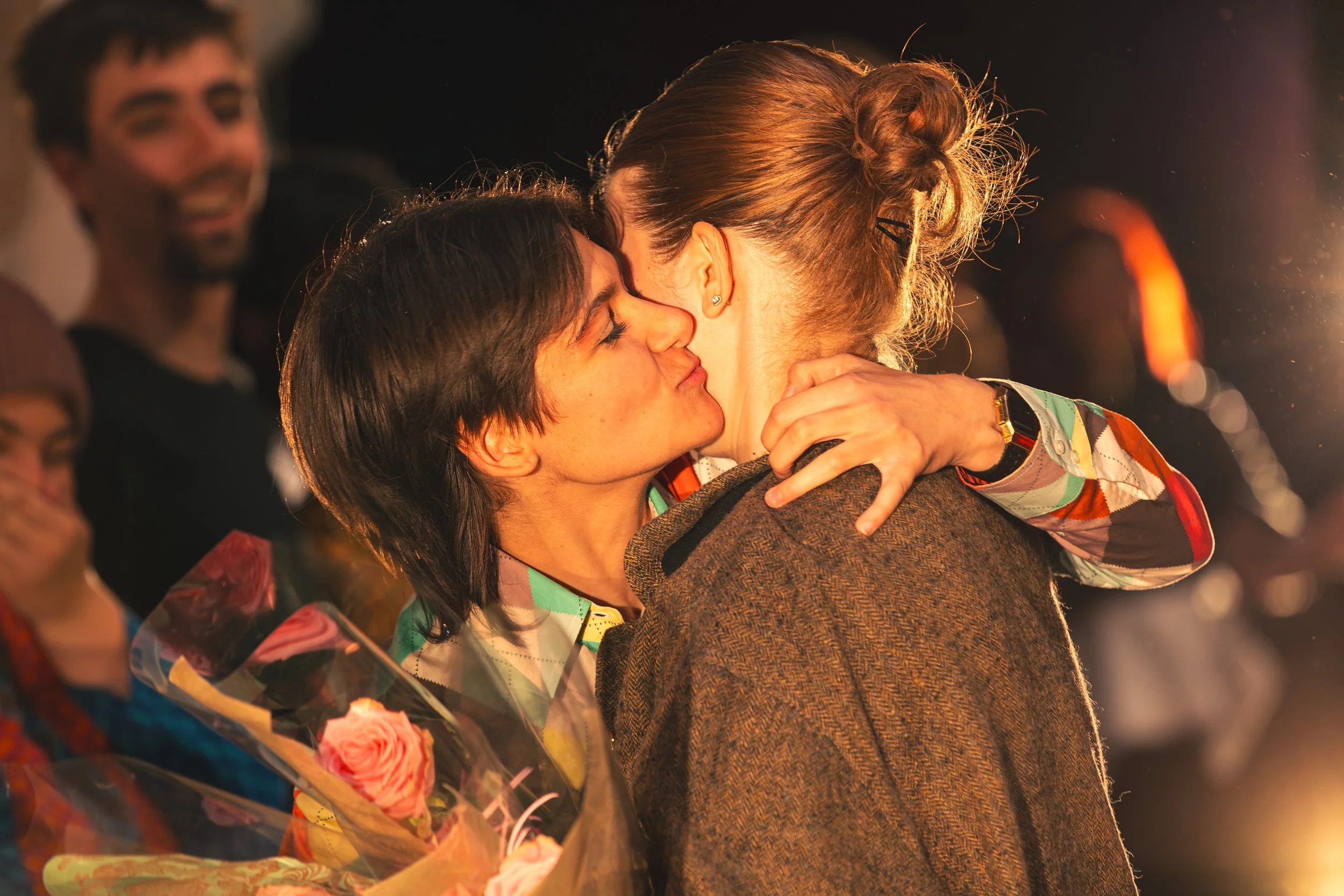 Deux femmes s'embrassent lors d'une célébration, une d'elles tient un bouquet de fleurs. En arrière-plan, d'autres personnes souriantes regardent.