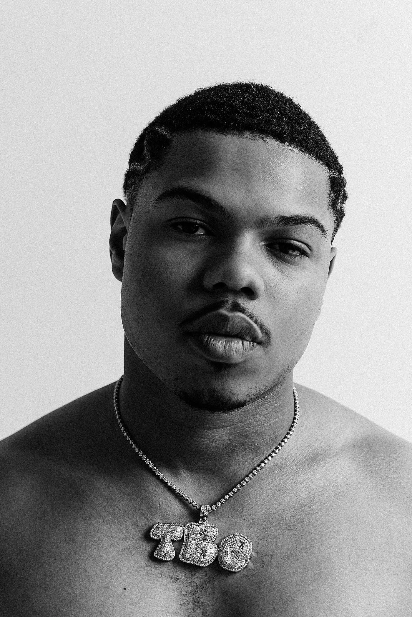Chicago rapper Taylor Bennet, wearing a chain necklace with large block letters 'TBG' encrusted with diamonds, against a plain light background.