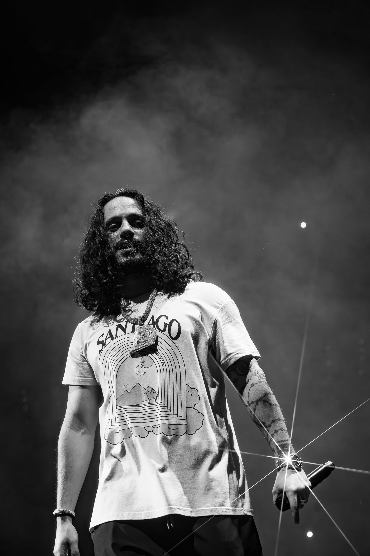Russ standing on stage, holding a microphone and wearing a T-shirt with a colorful graphic and the words 'SAN DIEGO'. The background is dark with some visible smoke or fog.