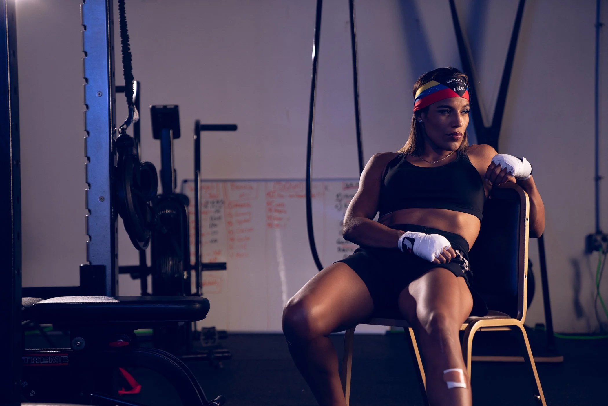 UFC Champion Julianna Pena sitting on a chair in a gym, wearing a black sports outfit, boxing gloves, and a colorful headband, appearing focused and tired.