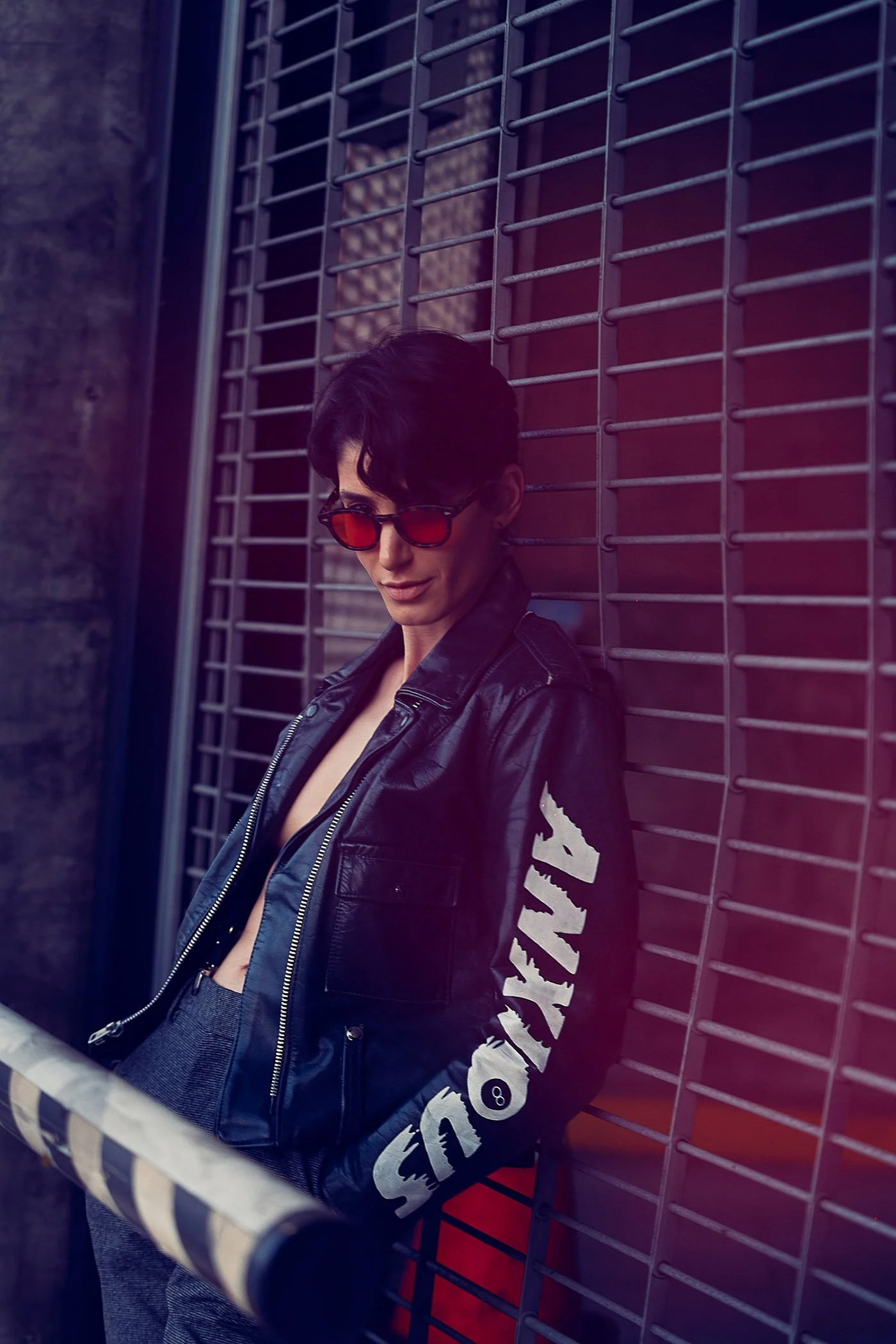 A person with short dark hair wearing a black leather jacket with white lettering on the sleeve, red-tinted sunglasses, and gray pants, standing against a metal grid wall with a dark brick background.