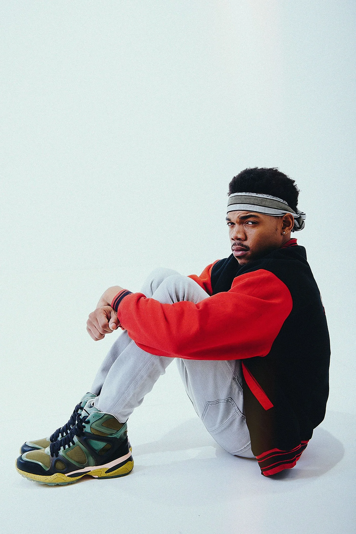 Chance the Rapper sitting on the floor, wearing a black and red jacket, light gray pants, and colorful sneakers, with a gray headband, against a plain light background.