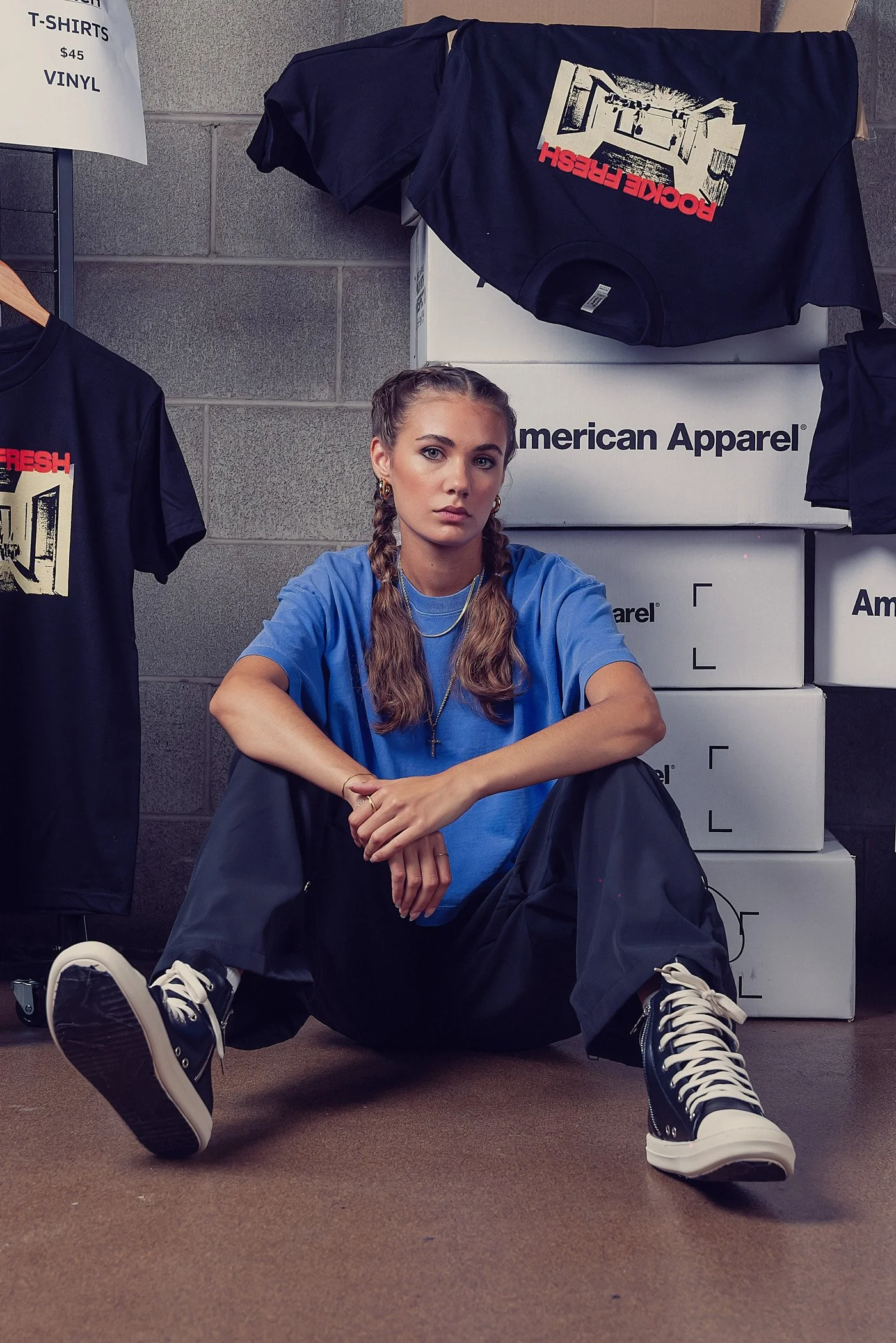 A young woman with braided hair, wearing a blue T-shirt, sitting on the floor with her legs apart. She is surrounded by boxes labeled American Apparel and T-shirts and is positioned in front of a stack of boxes and clothing racks, with serious facial